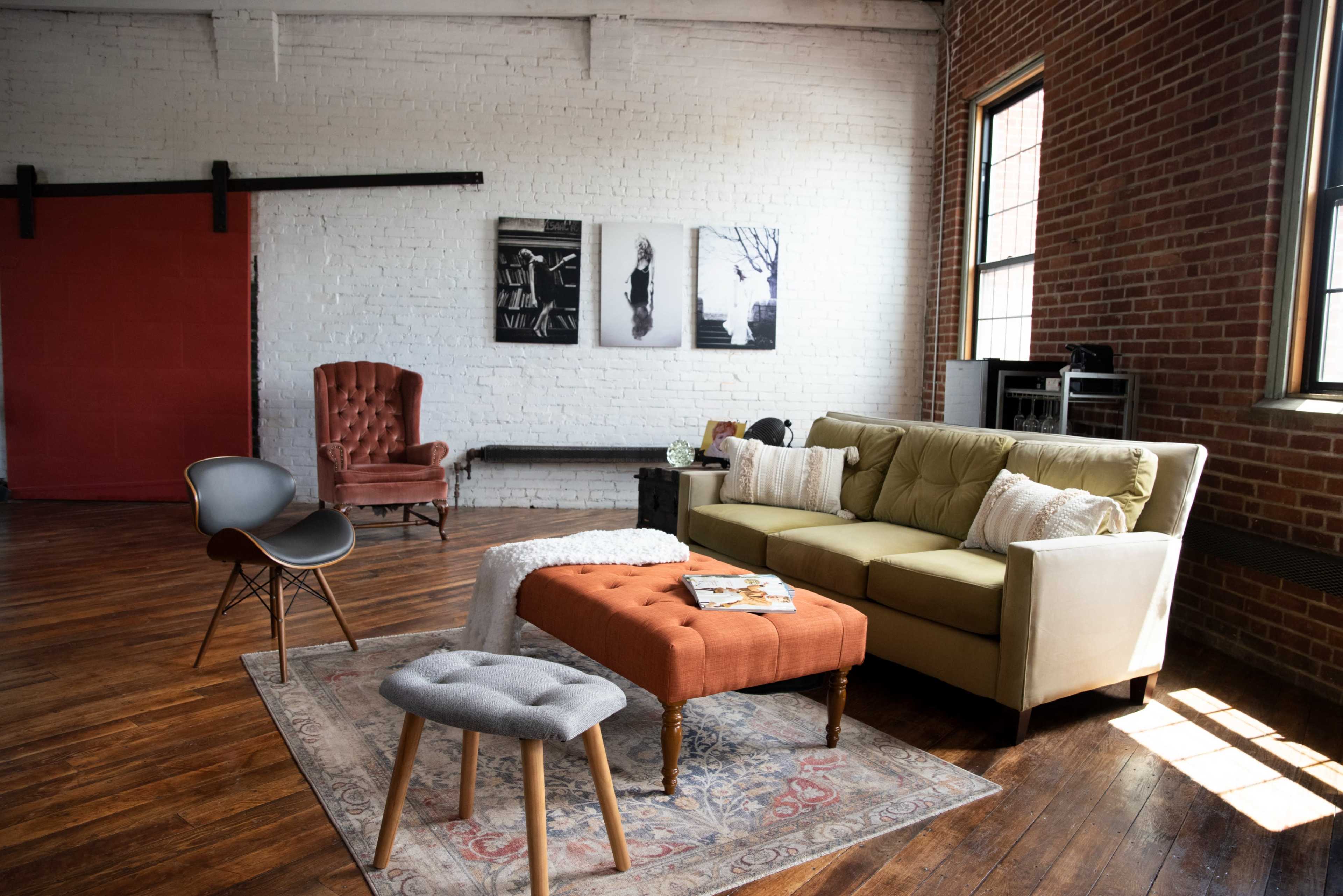 A cozy living room featuring a green sofa, an orange upholstered ottoman, a gray stool, and a burgundy armchair, with exposed brick walls and wooden floors.