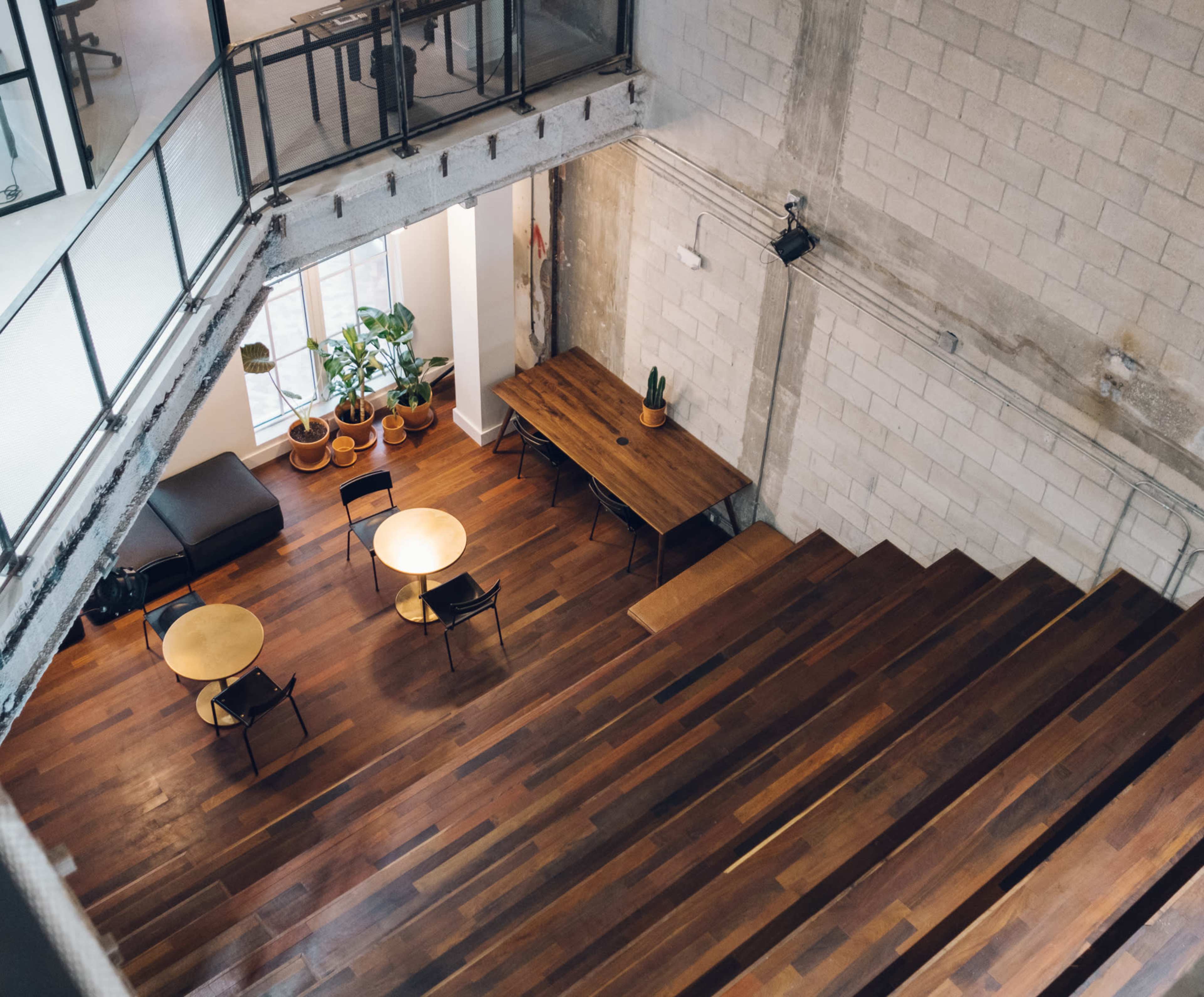The image shows a spacious interior with wooden flooring, featuring multiple tables and chairs, and large windows allowing natural light.