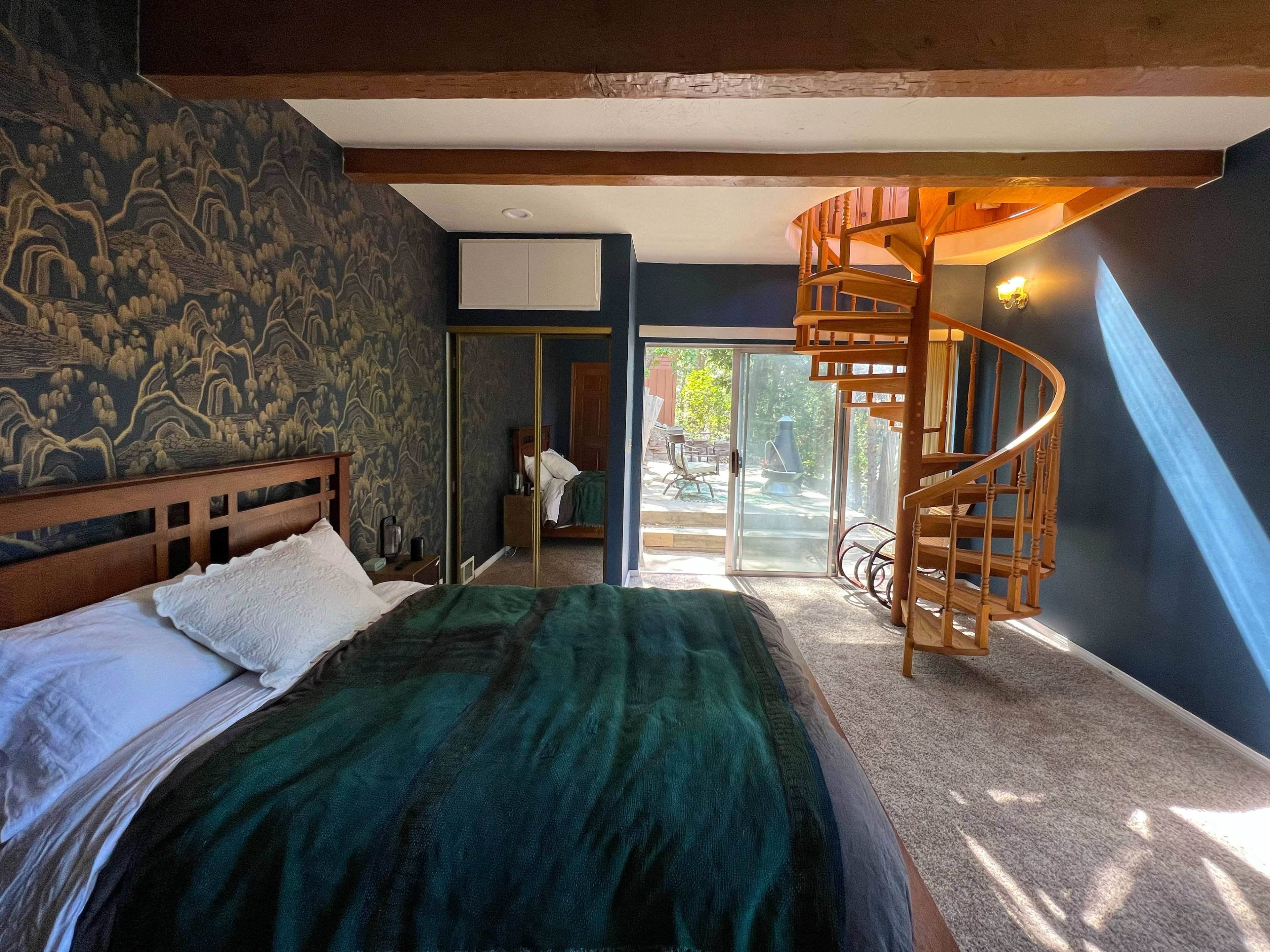 A bedroom with a wooden bed, a patterned accent wall, a spiral staircase, and a sliding glass door leading to an outdoor area.