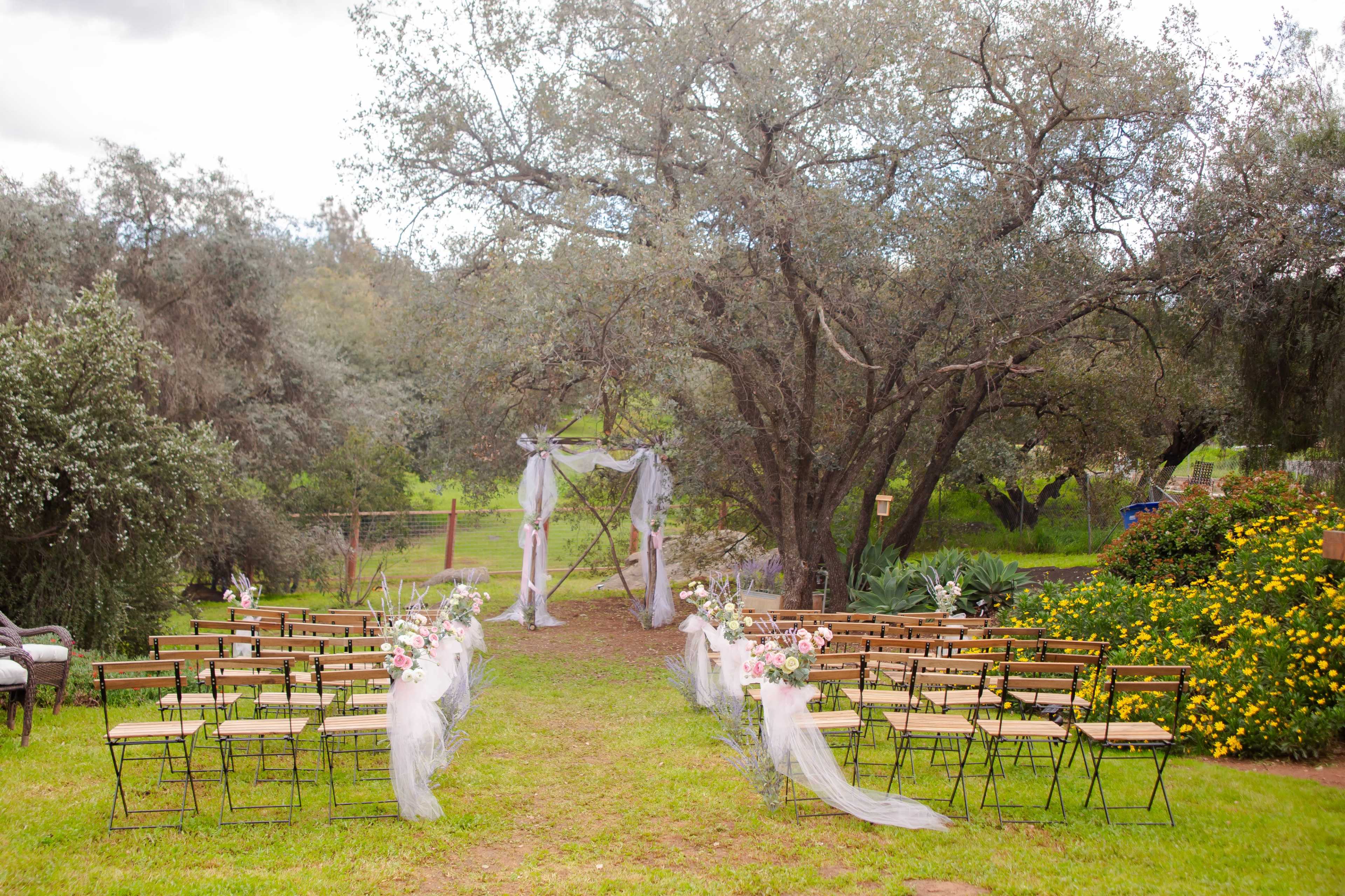 A wedding ceremony setup features rows of chairs arranged on a grassy area beneath large trees, with floral decorations and draped fabric framing an altar.