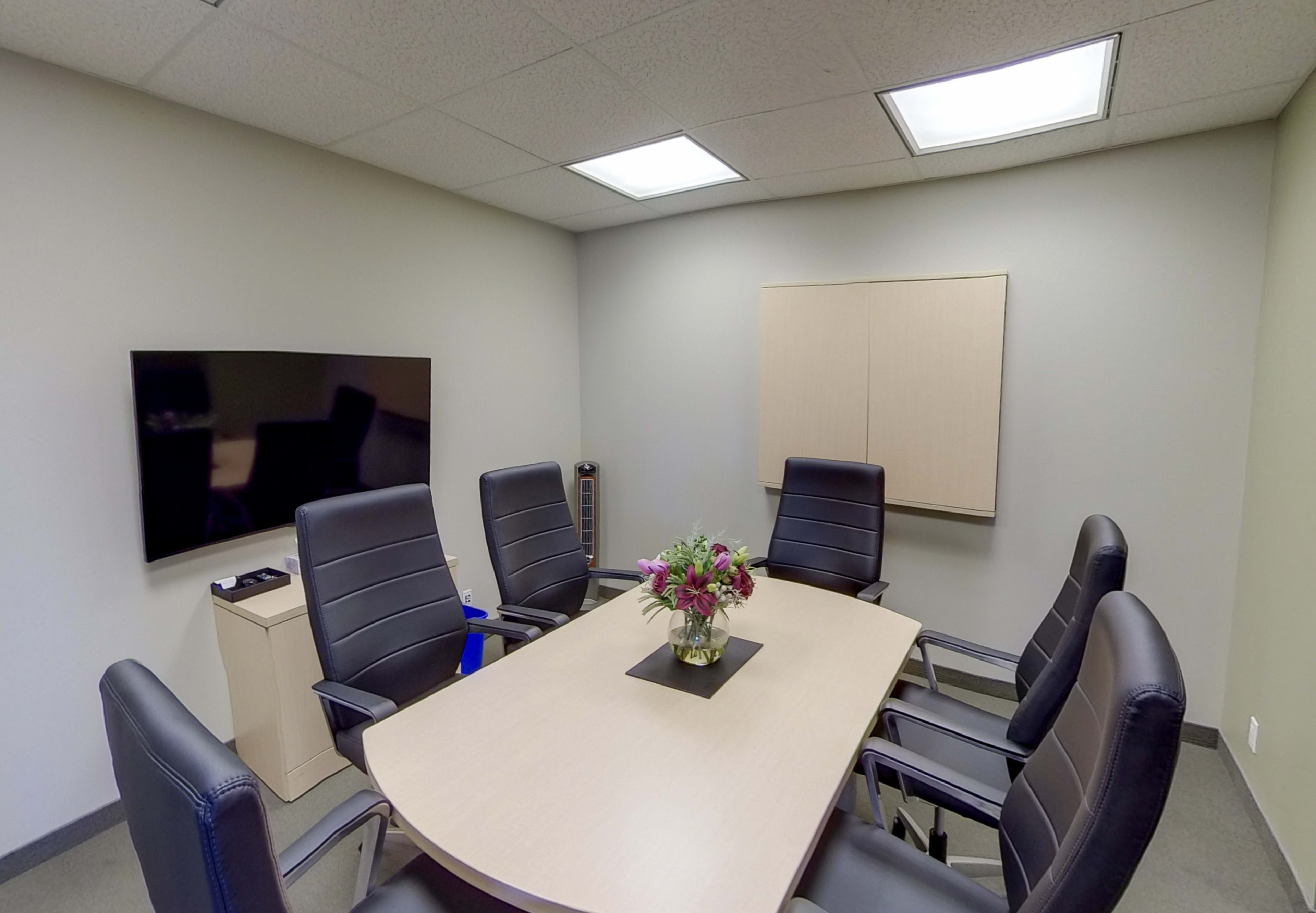 A conference room features a large table surrounded by six black chairs, with a television on the wall and a vase of flowers in the center.