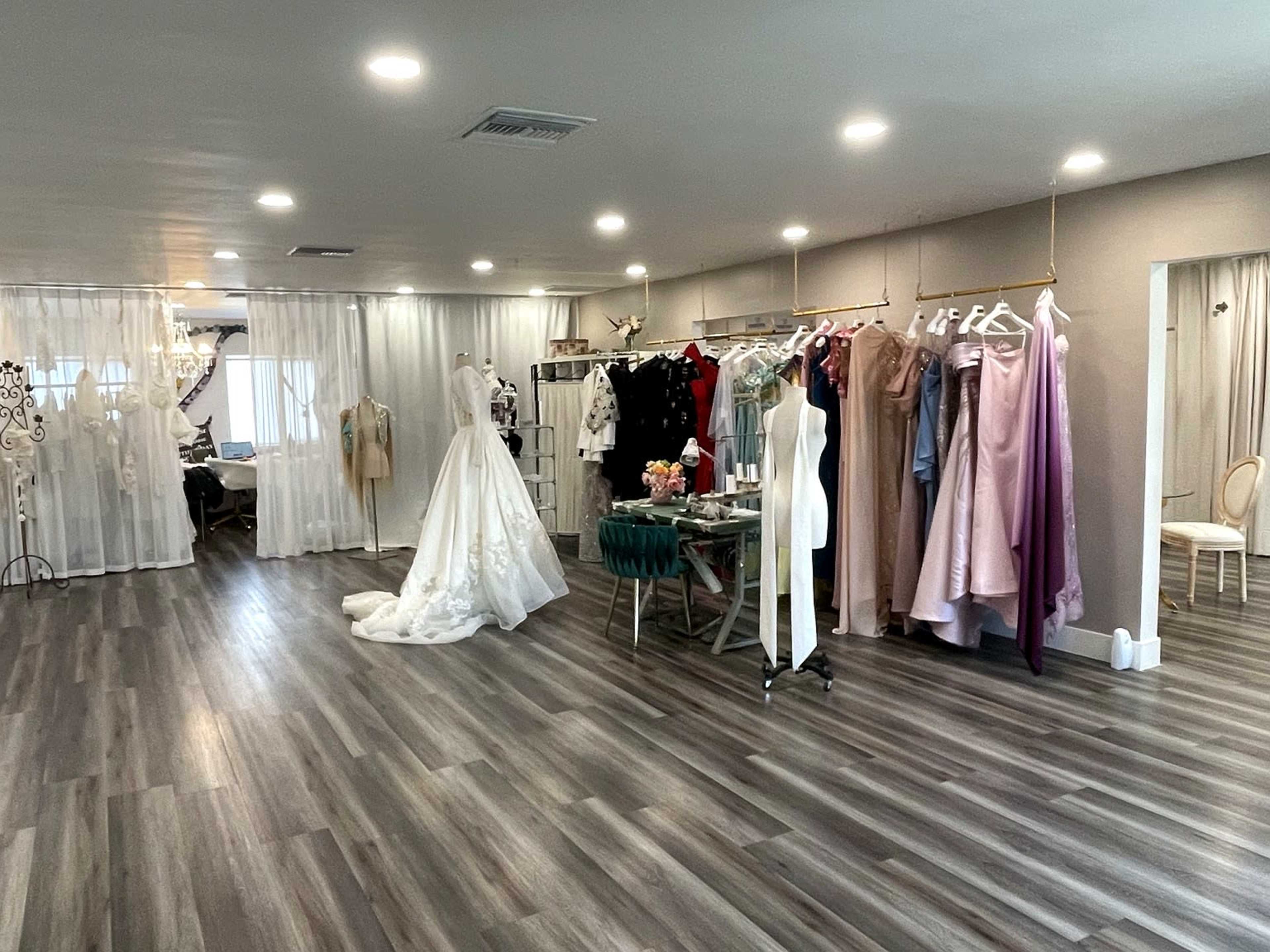 The image shows a spacious boutique interior with a variety of dresses displayed on racks, a sewing area in the background, and natural light coming through sheer curtains.