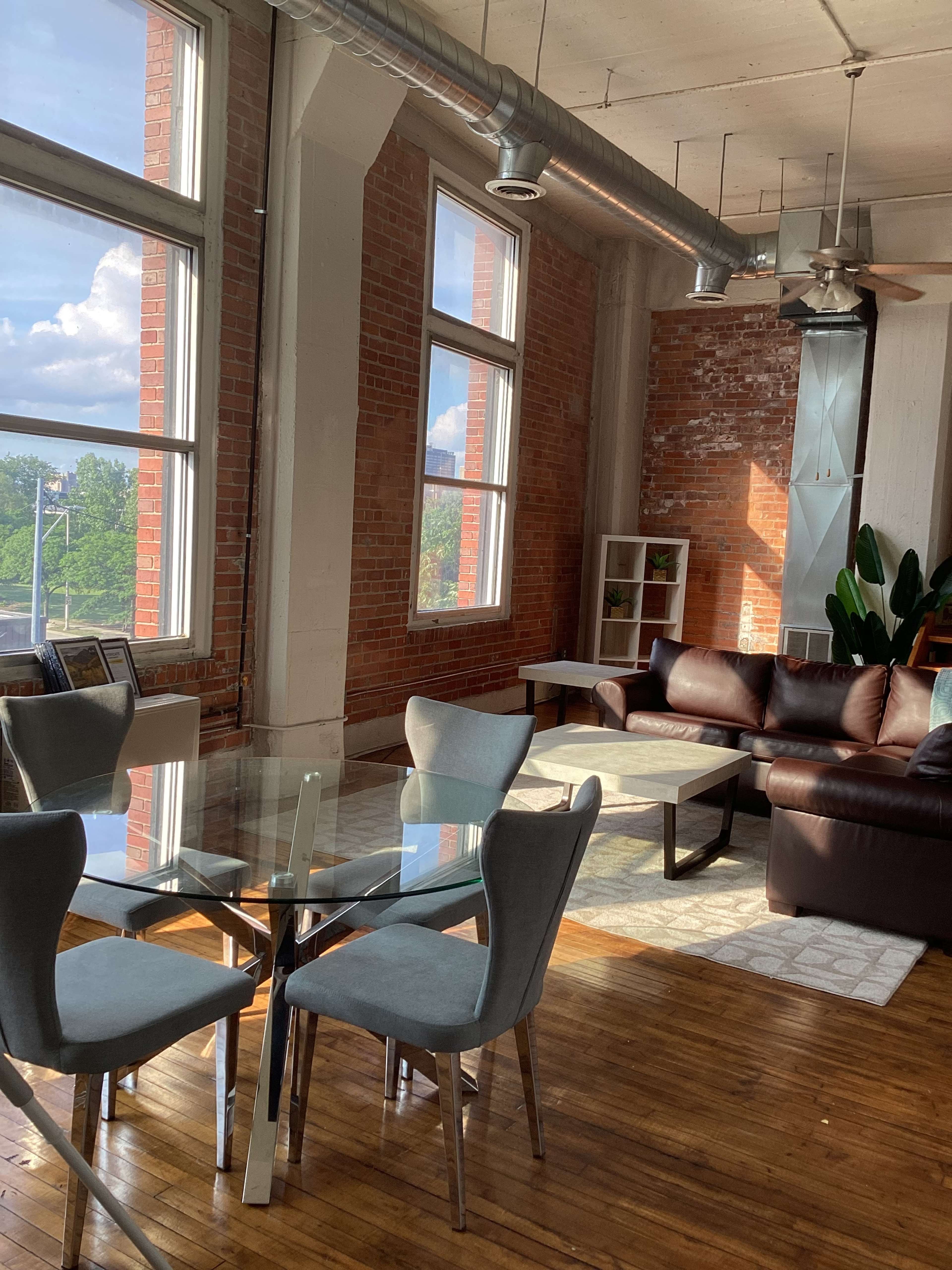 The image shows a bright, modern living space featuring large windows, a glass dining table with chairs, a brown leather sofa, and a potted plant.