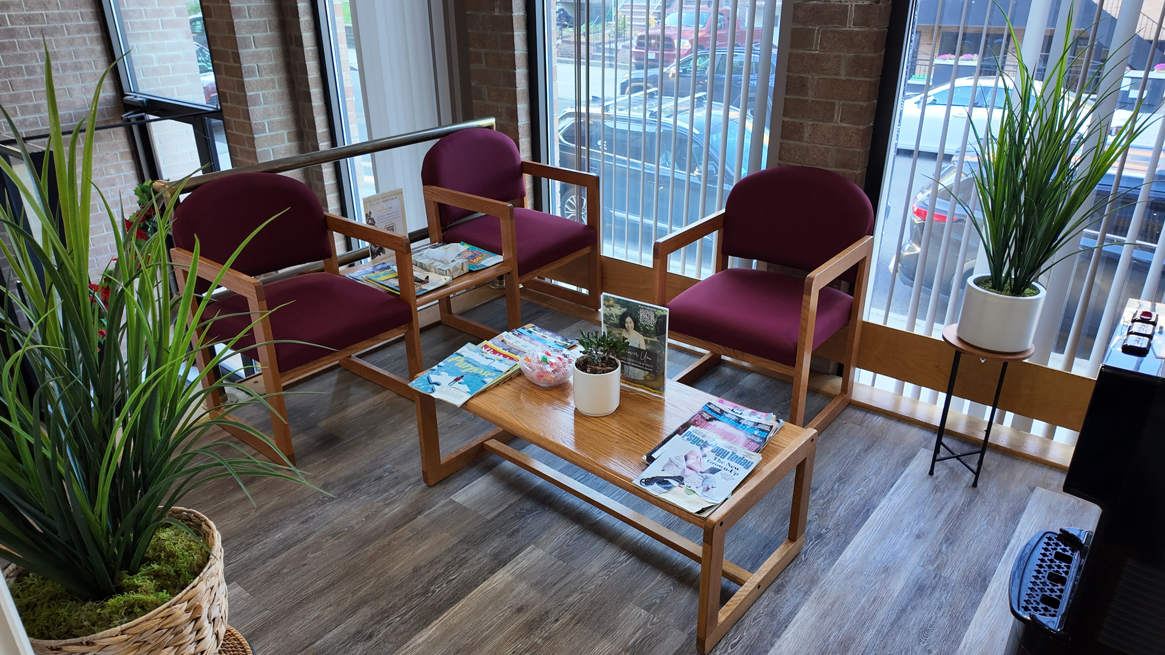 A waiting area features two maroon chairs arranged around a wooden table, with magazines on the table and potted plants in the corners.