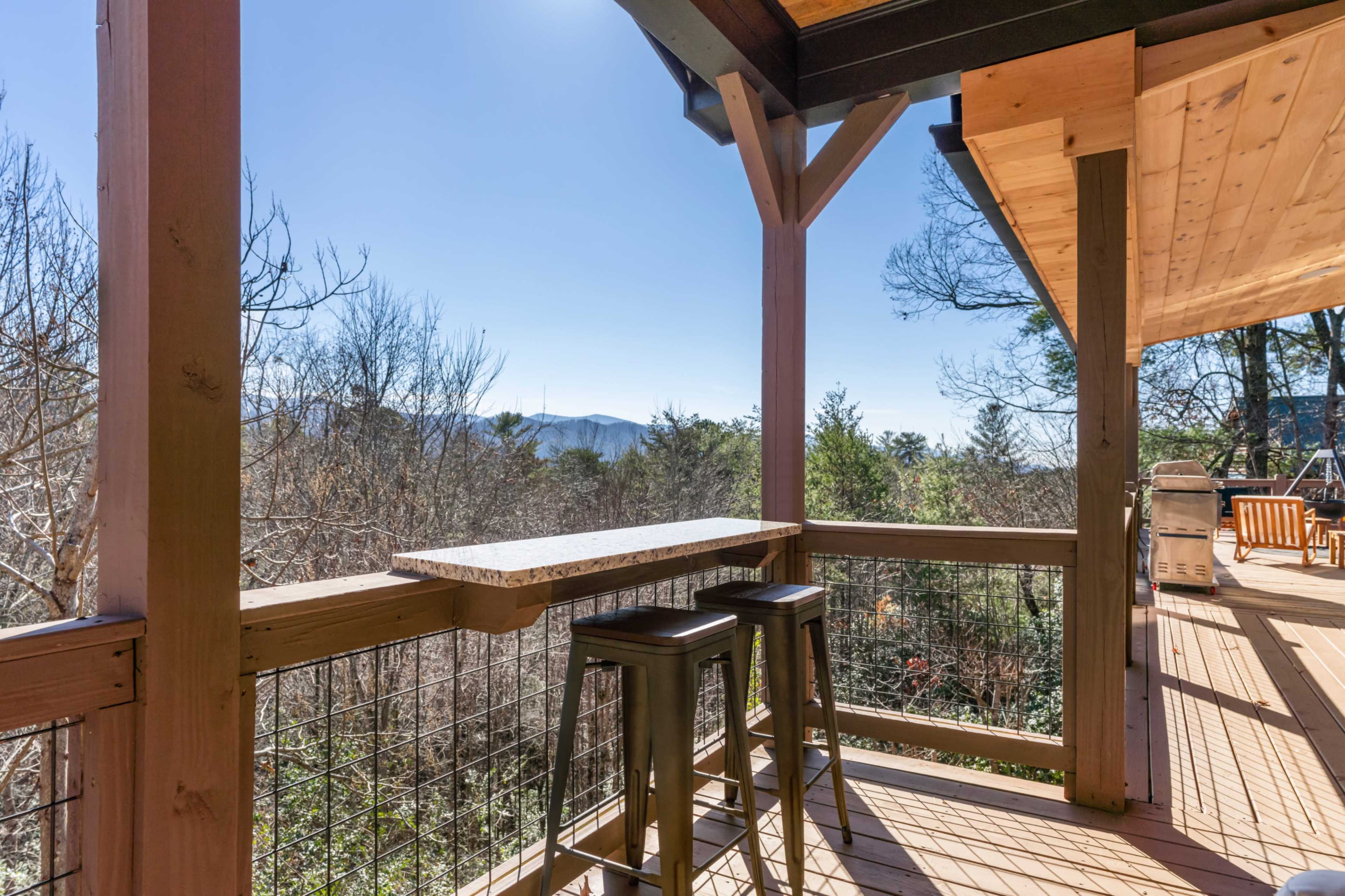 A wooden deck with two bar stools overlooking a landscape of trees and mountains under a clear blue sky.