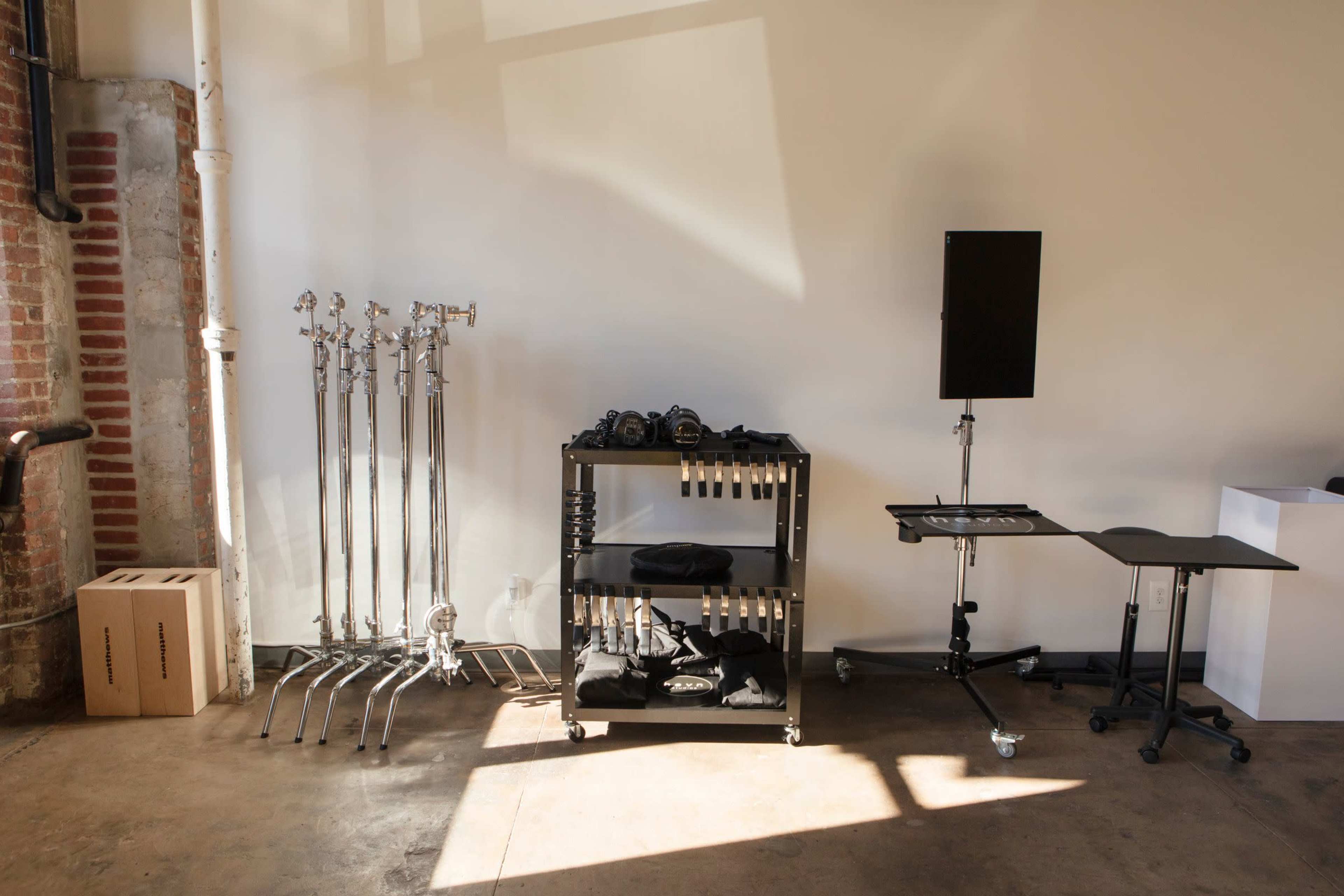 The image shows a well-lit interior space featuring chrome light stands, a black utility cart with equipment, and a small workspace with a table and a monitor.