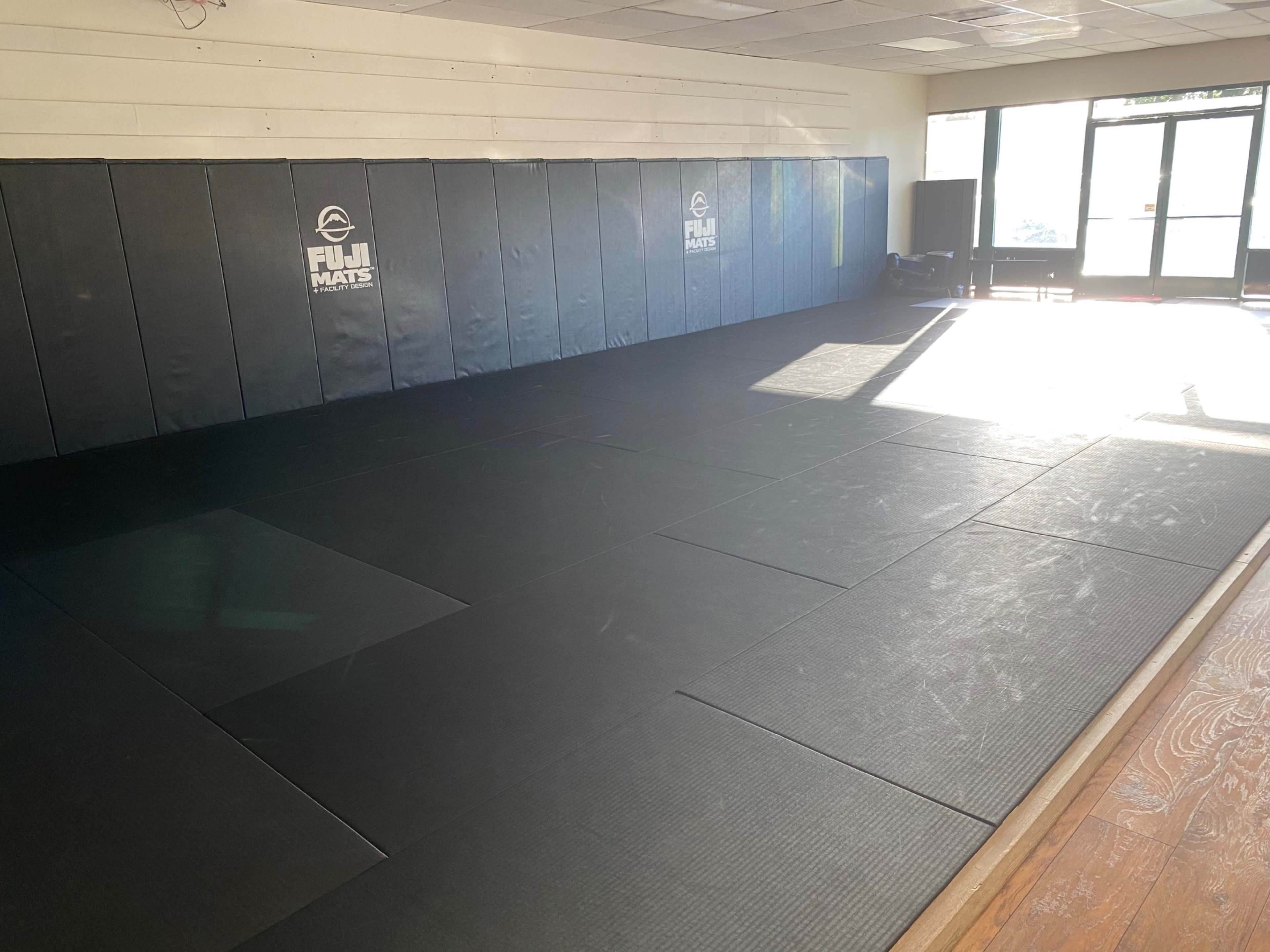 A large, empty martial arts training room with black mats and padded walls, illuminated by natural light from large windows.