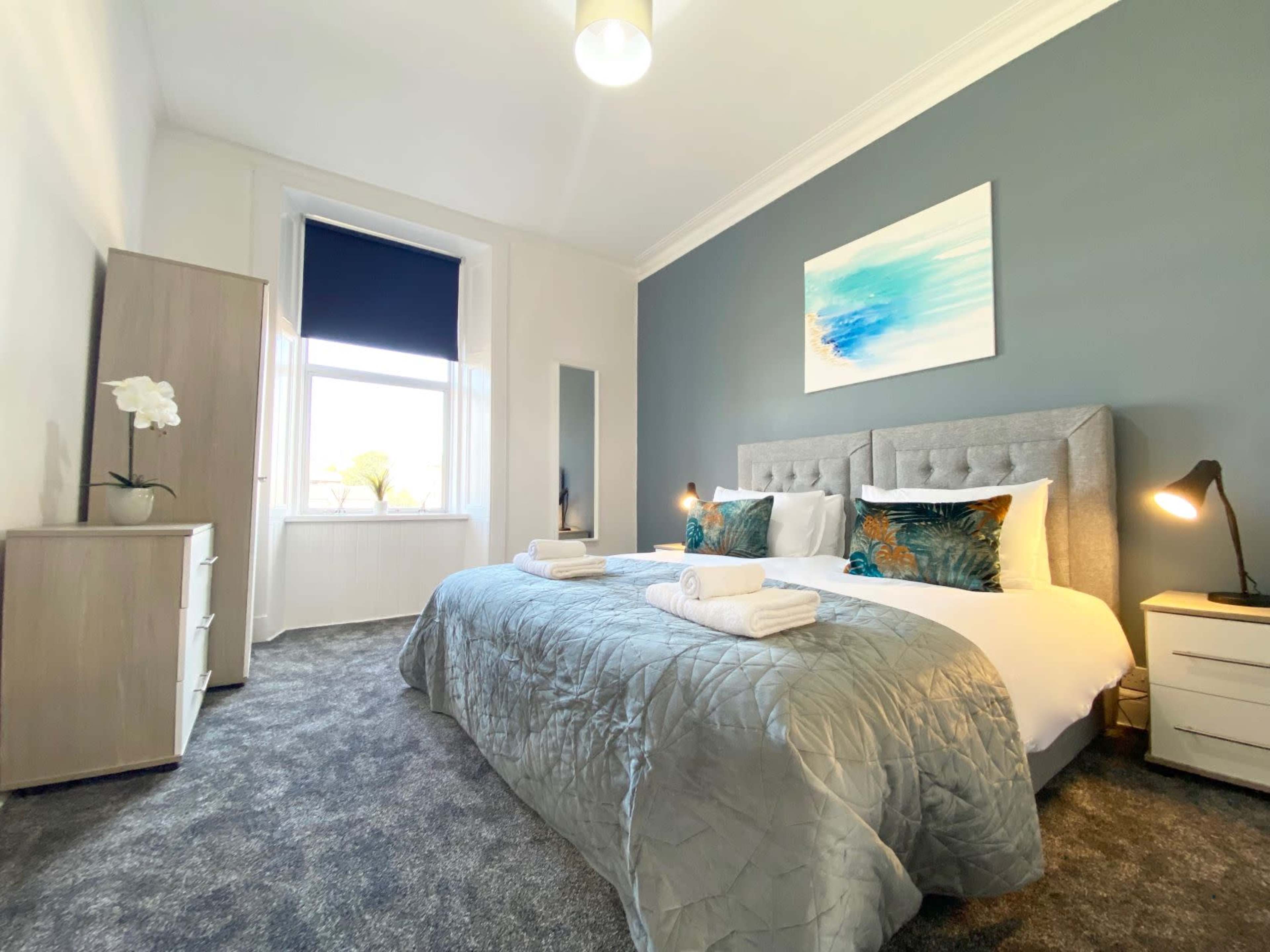 A spacious bedroom features a large bed with decorative pillows, a bedside table with a lamp, a dresser, and a window with a dark roller shade.