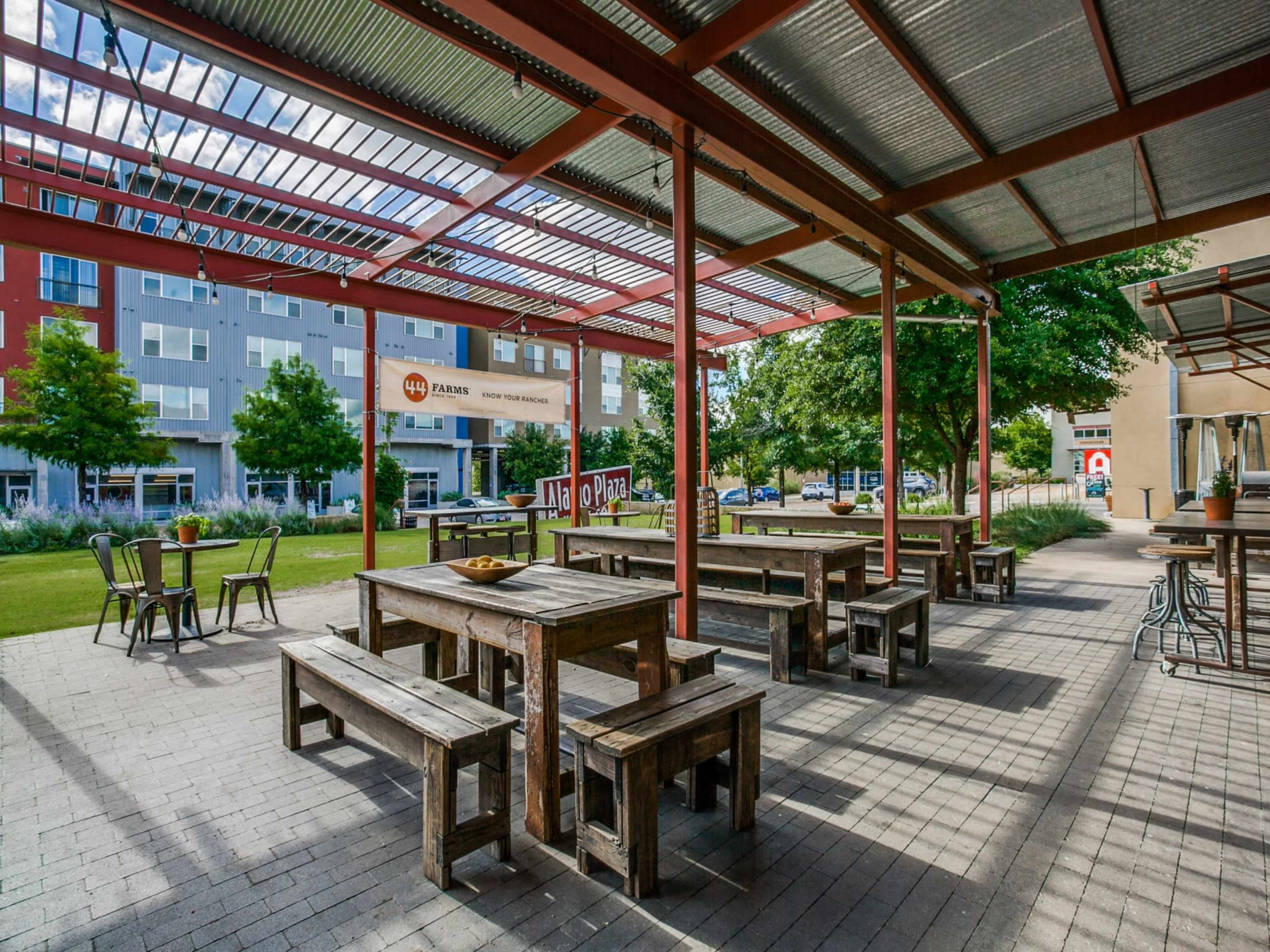 An outdoor seating area with wooden benches and tables under a metal roof, surrounded by greenery and modern buildings.