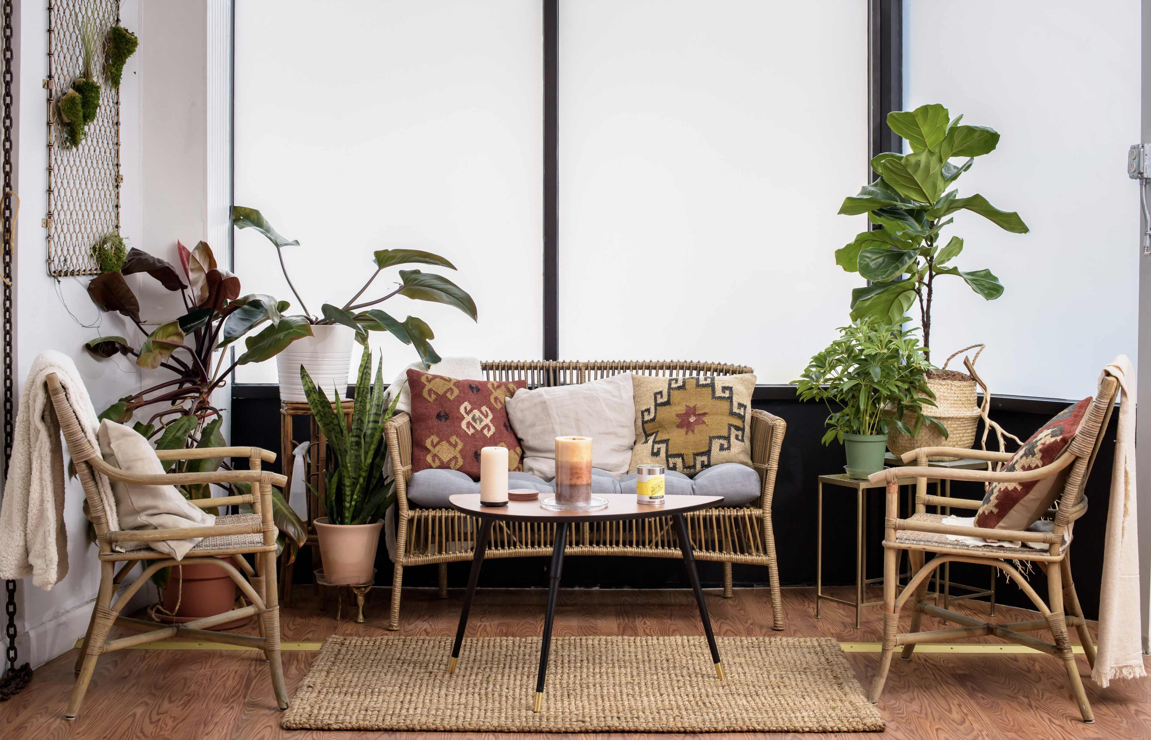 A cozy indoor seating area features a wicker sofa and two matching chairs surrounded by various potted plants and a small table with candles.