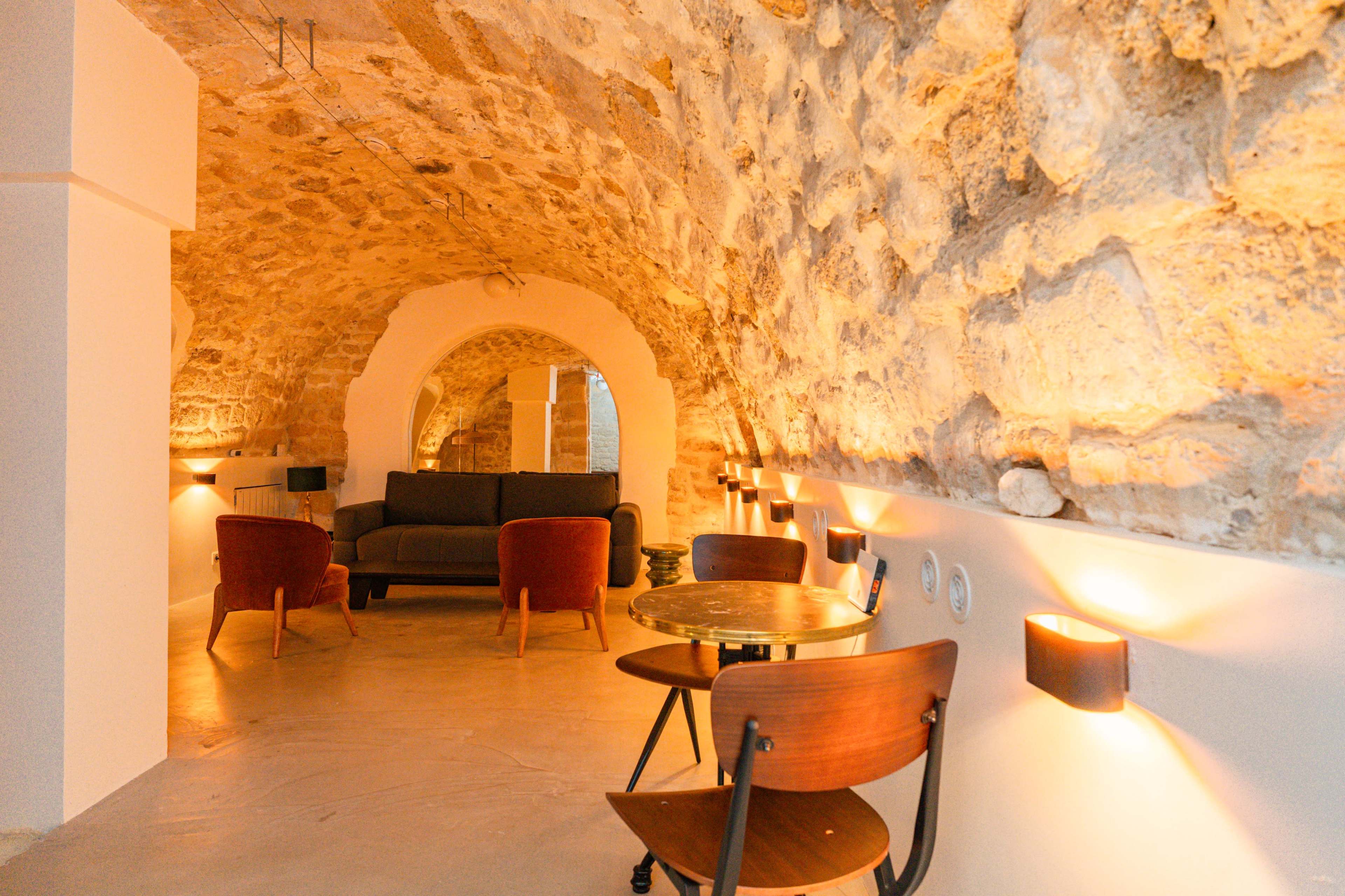 The image shows a cozy interior space featuring a stone arch ceiling, a sofa, a small round table, and two chairs, illuminated by warm wall sconces.