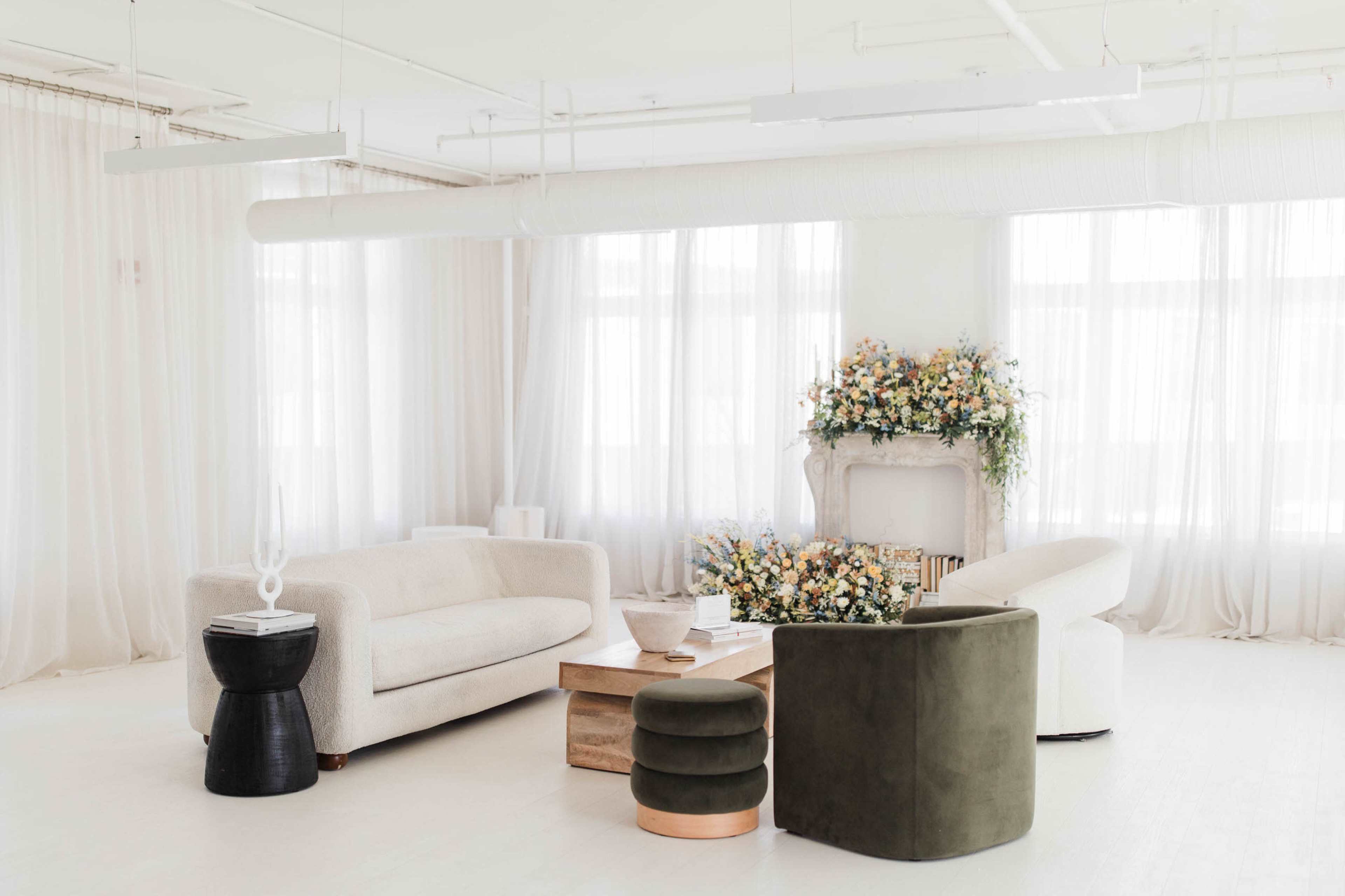 A bright, modern living room features a light-colored sofa, a circular green chair, a wooden coffee table, and a floral arrangement on a decorative mantel, all set against sheer white curtains.