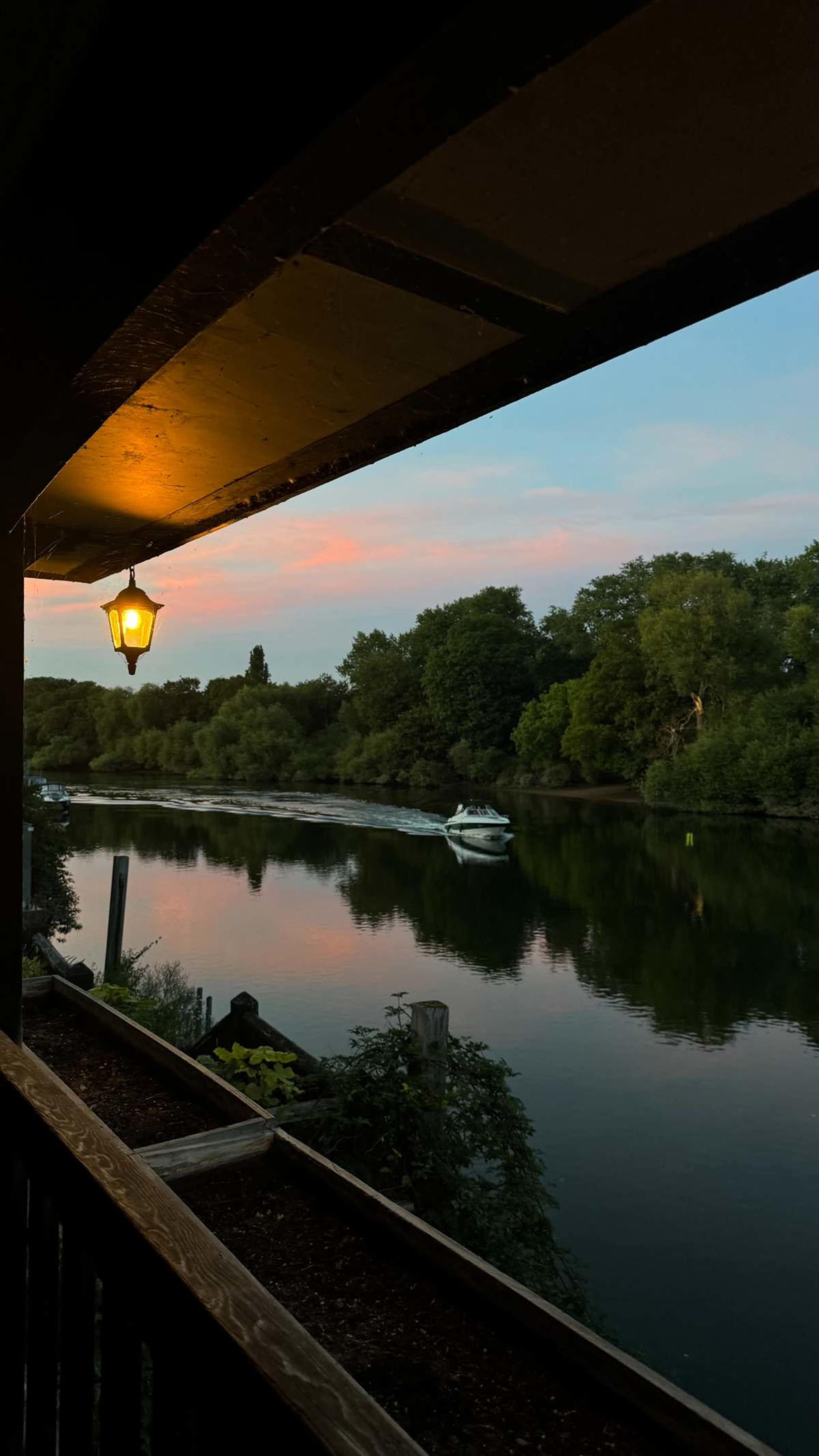 A lantern hangs over a peaceful river scene, where a boat glides through the water surrounded by lush greenery and a colorful sky at dusk.