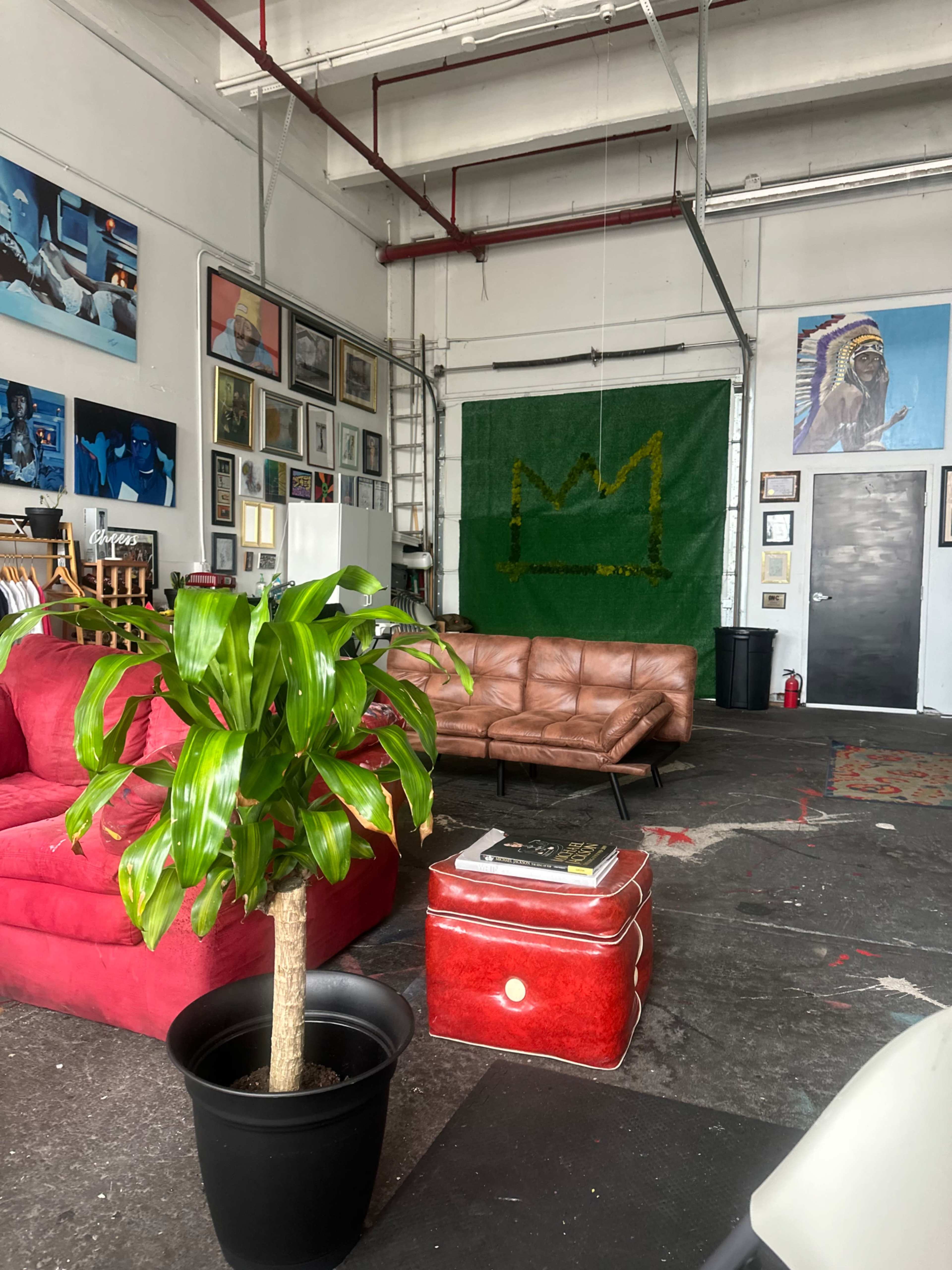 The image shows an industrial-style interior with a brown leather sofa, a red armchair, a potted plant, and various artworks on the walls.