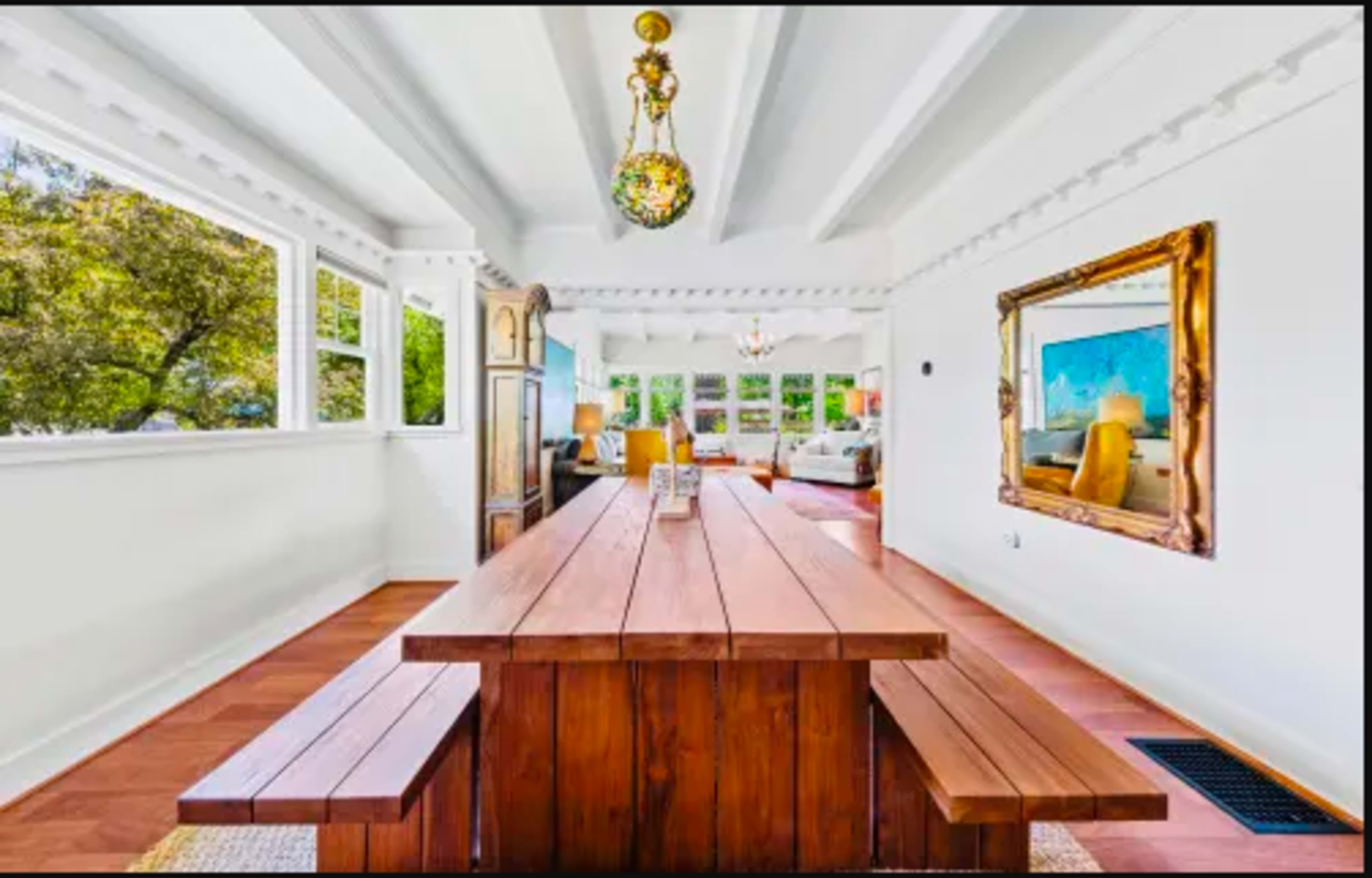 A bright, open dining area with a long wooden table and benches, featuring large windows that look out onto greenery.