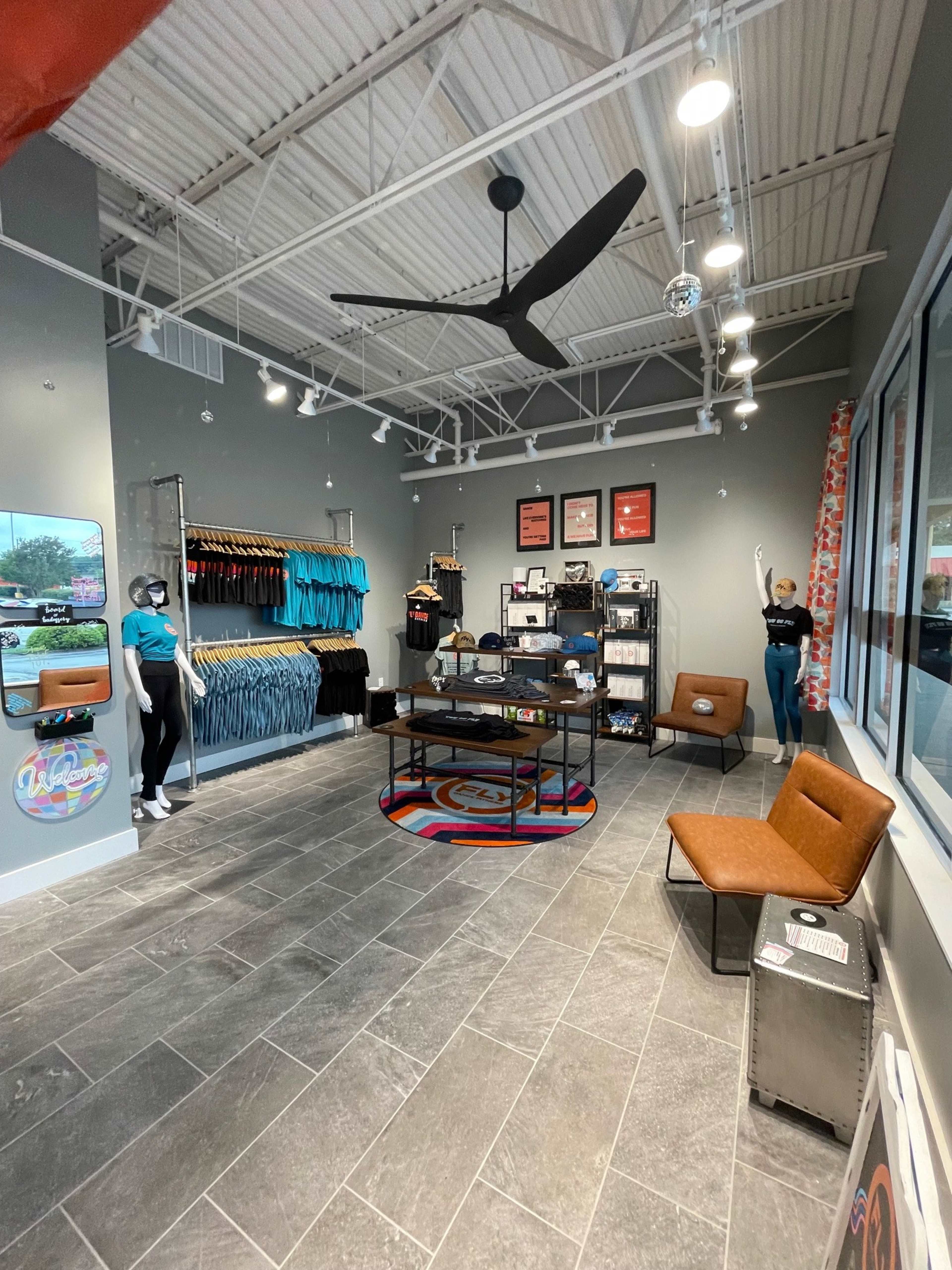 The image shows a retail store interior featuring a display of clothing on racks, a central table with merchandise, and a seating area, all under bright lighting.