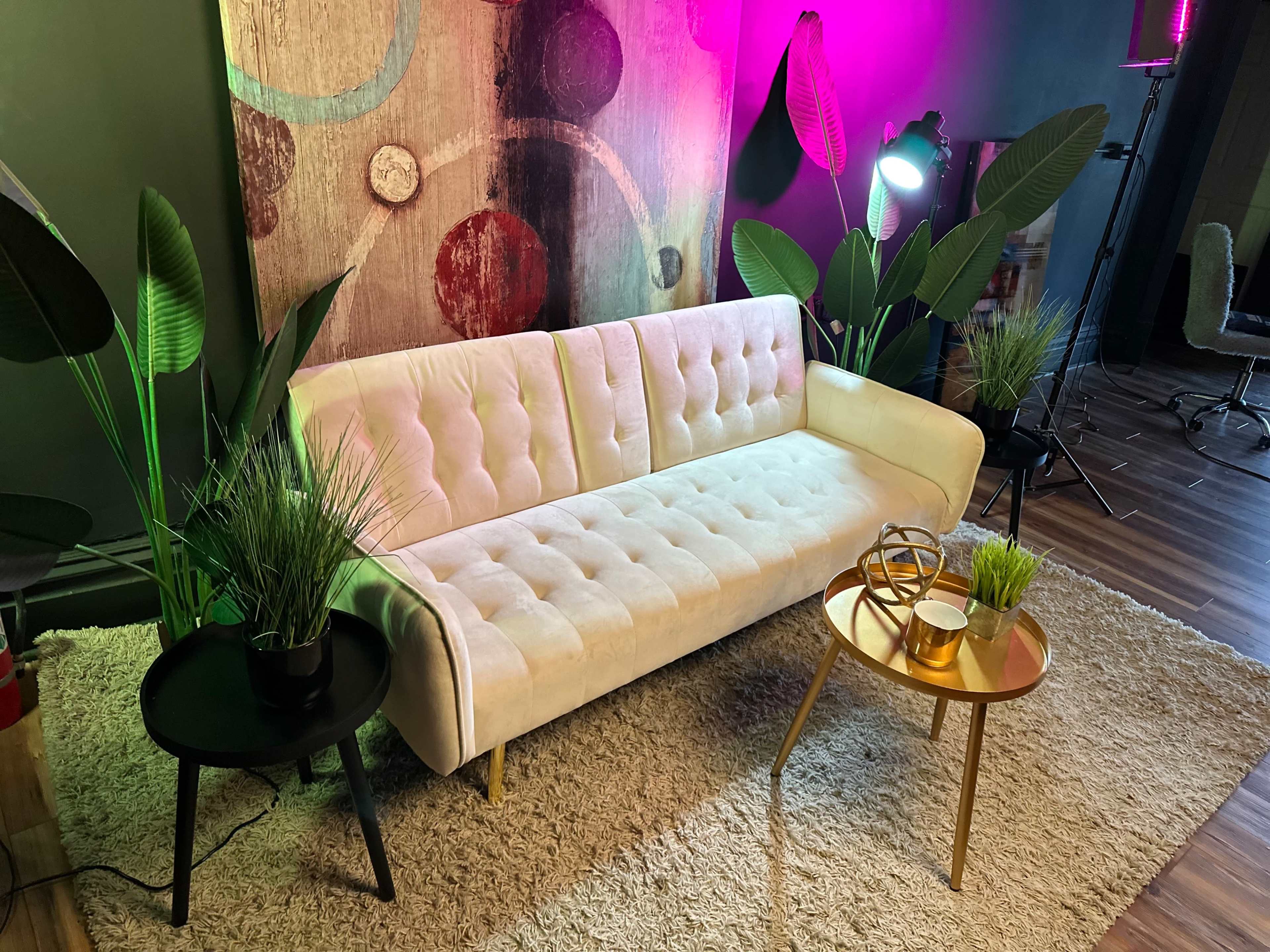 A modern living room scene featuring a beige tufted sofa surrounded by indoor plants, a small round table with decorative items, and vibrant lighting.