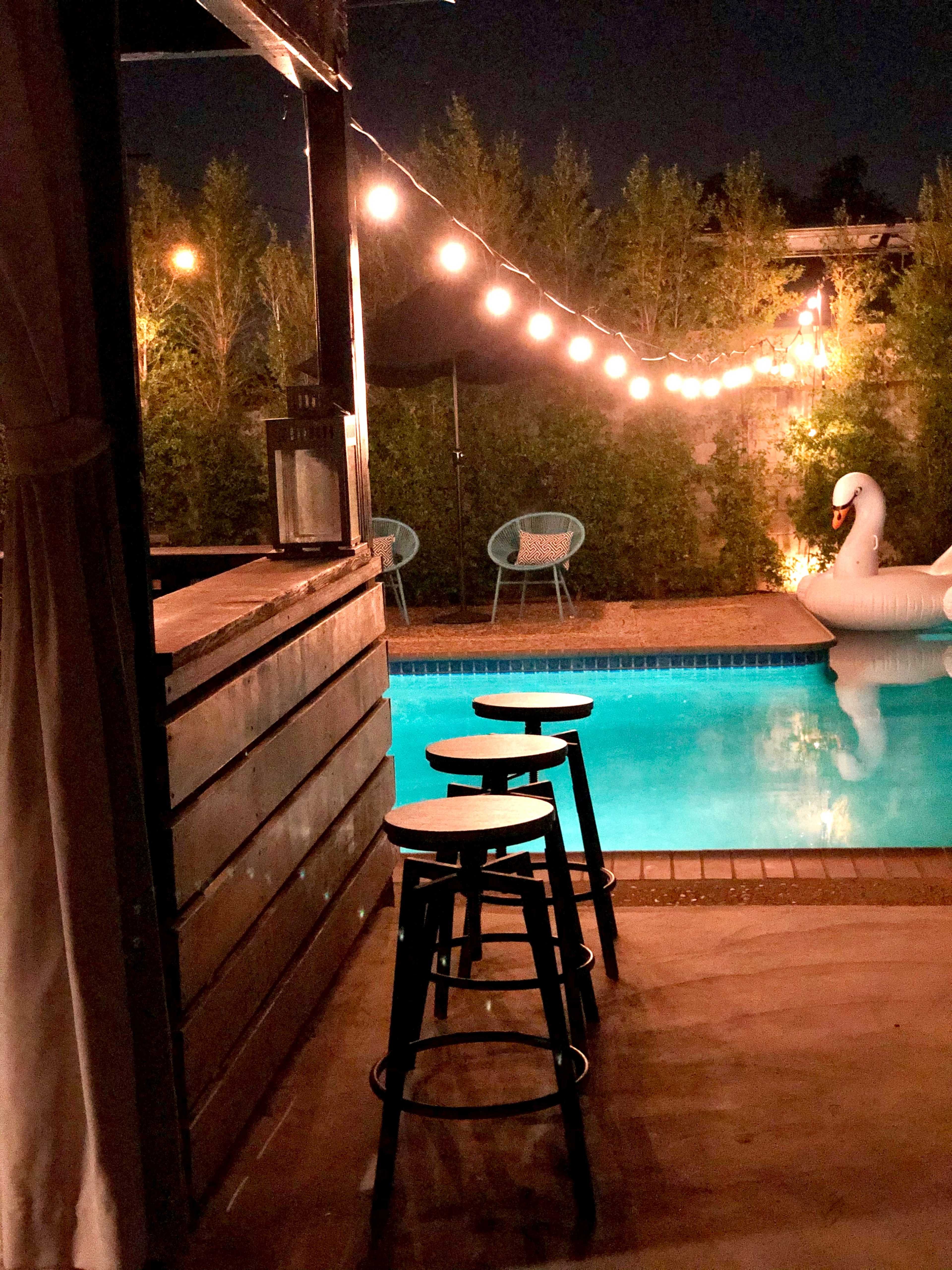 The image shows a pool area at night with a bar, three stools, and string lights illuminating the space beside a large inflatable swan.