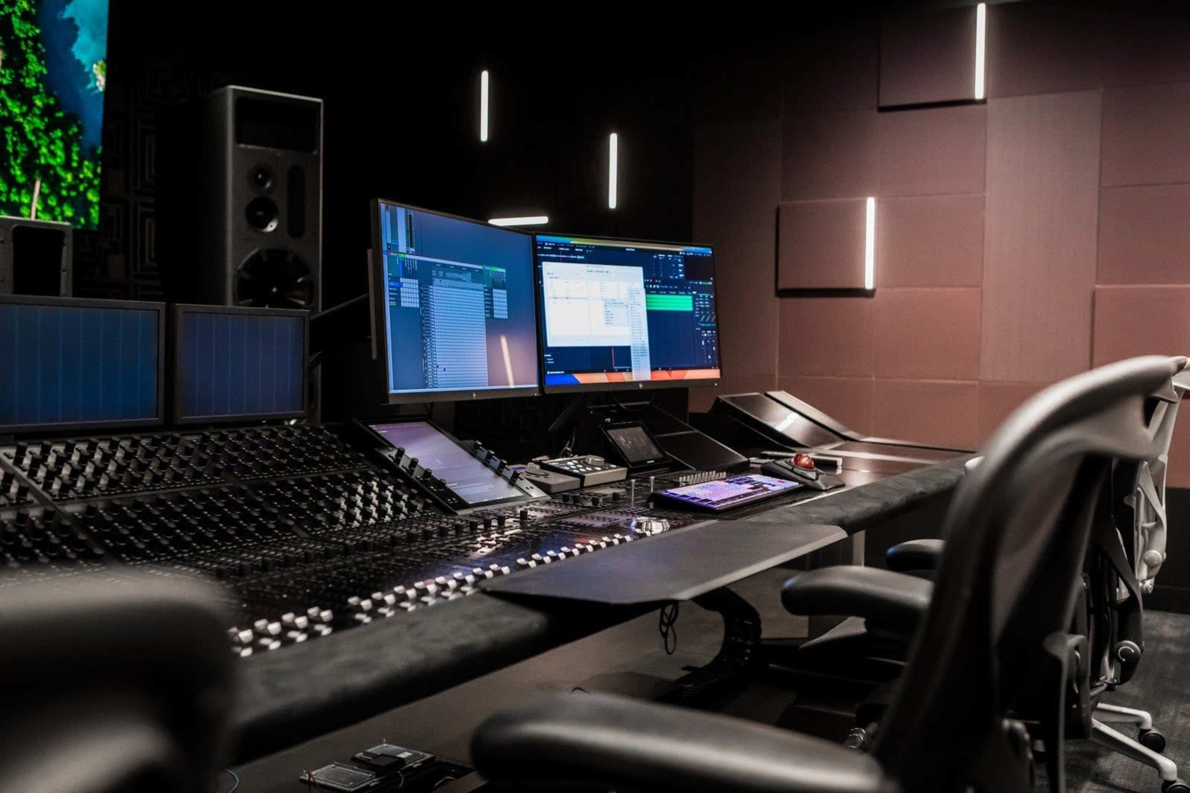 The image shows a modern audio mixing studio featuring a large control desk with various knobs and sliders, multiple monitors, and a sound system setup against a dark backdrop.