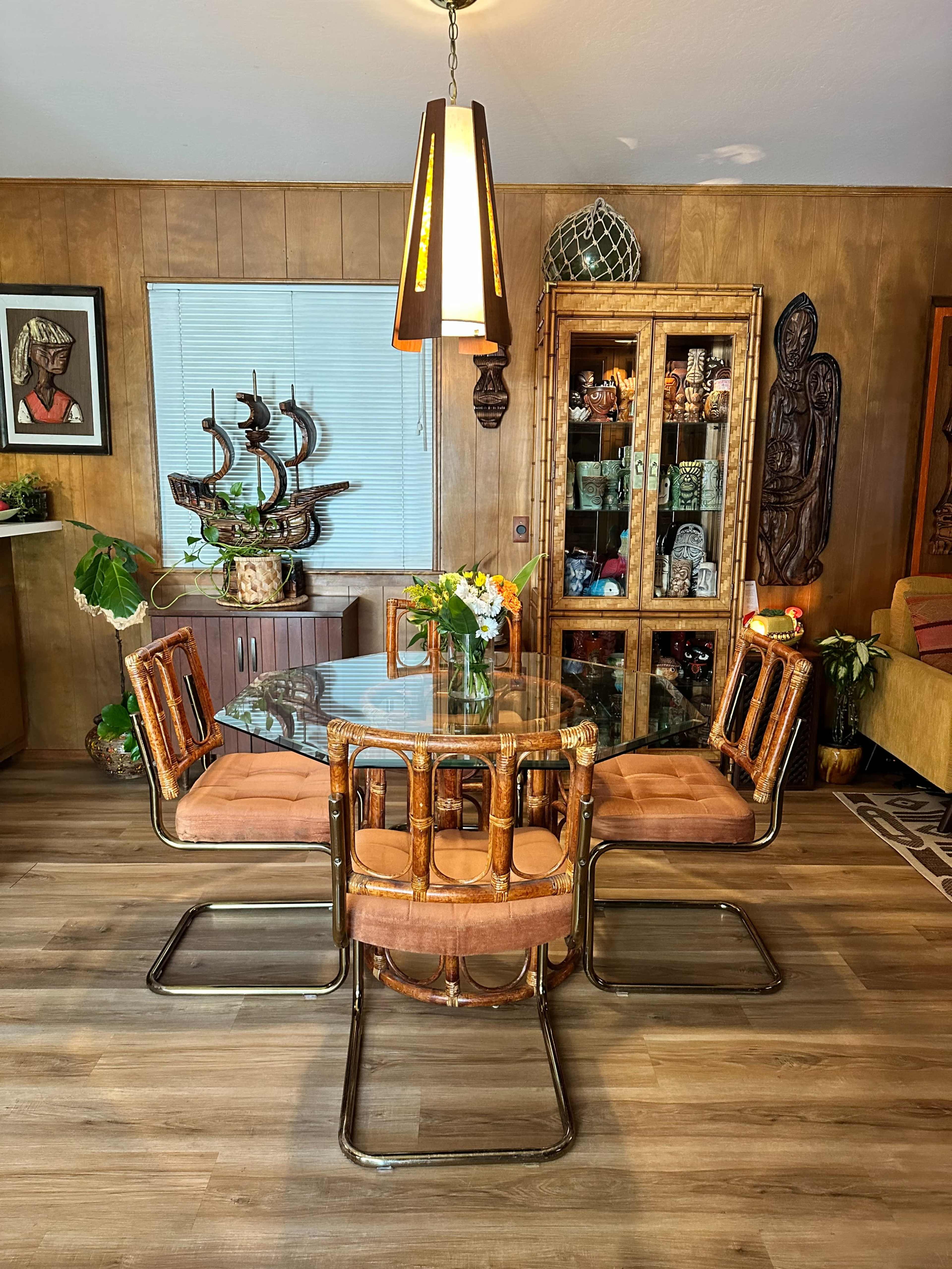 A glass-top dining table surrounded by four bamboo chairs is illuminated by a modern pendant light in a wooden-paneled room featuring decorative shelves and artwork.