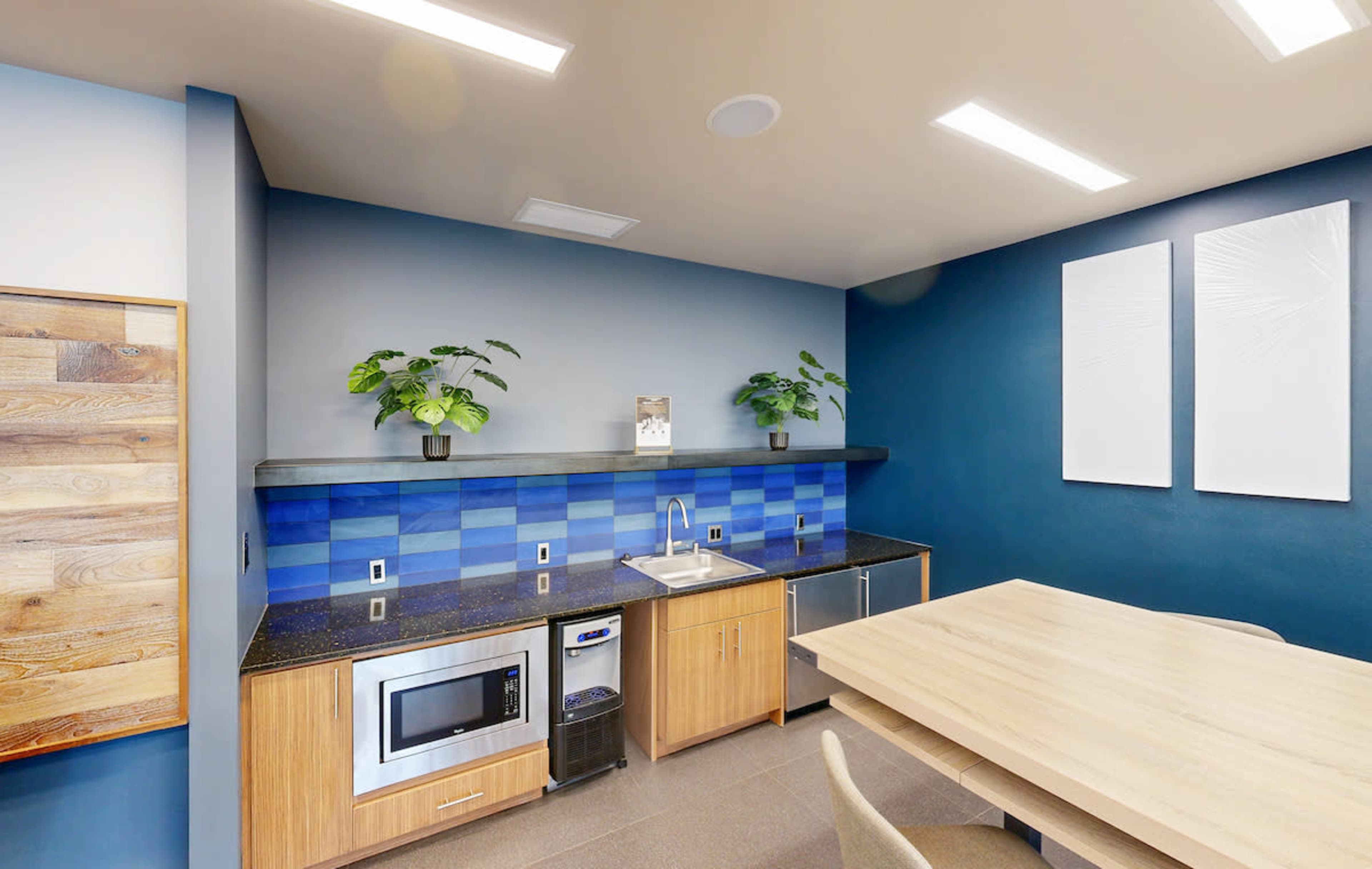 The image shows a modern kitchen area featuring a blue tiled backsplash, a sink, a microwave, and a wooden dining table with chairs.