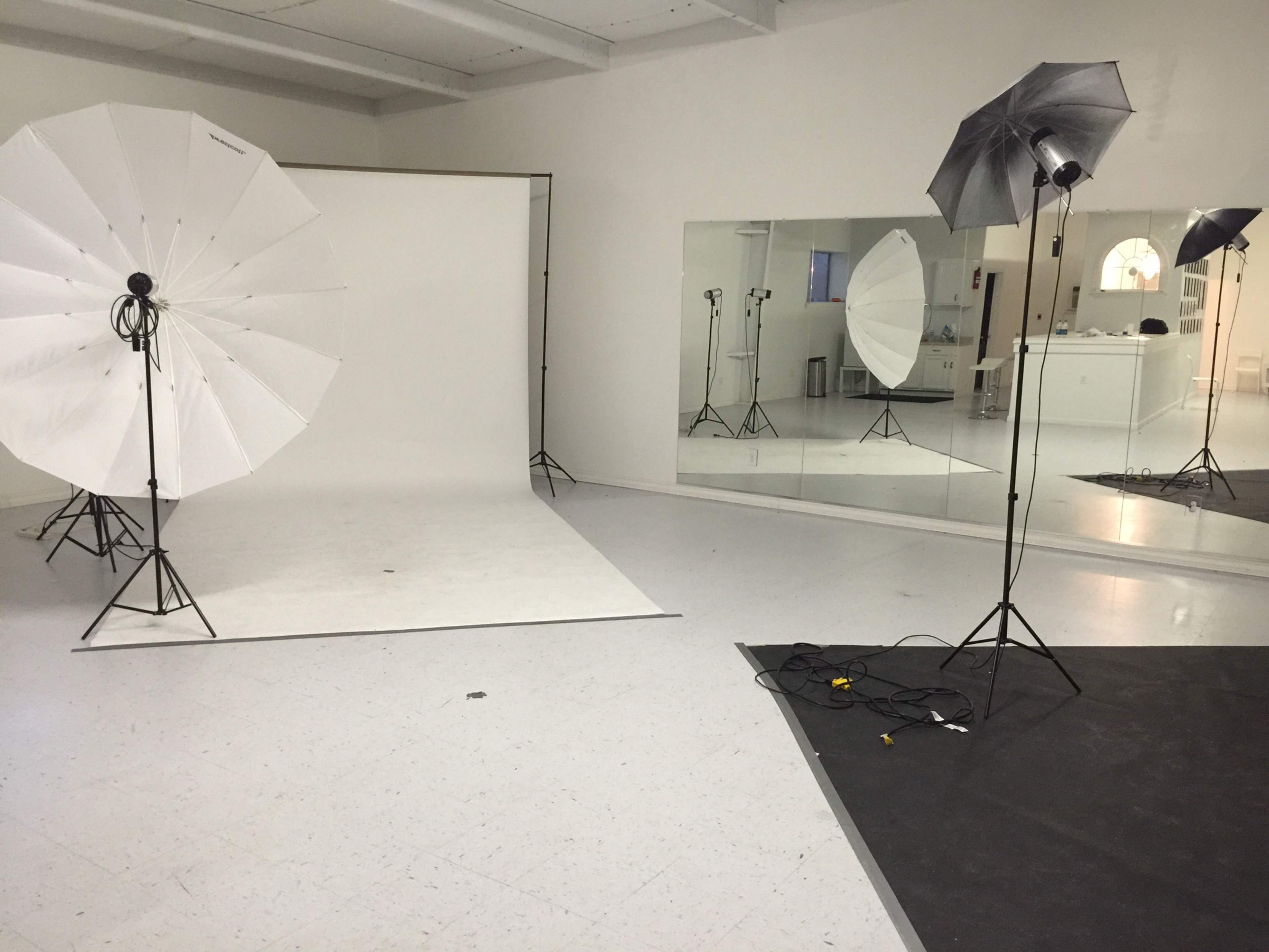 The image shows a photography studio with two large white umbrella lights, a backdrop, and mirrors reflecting the setup.