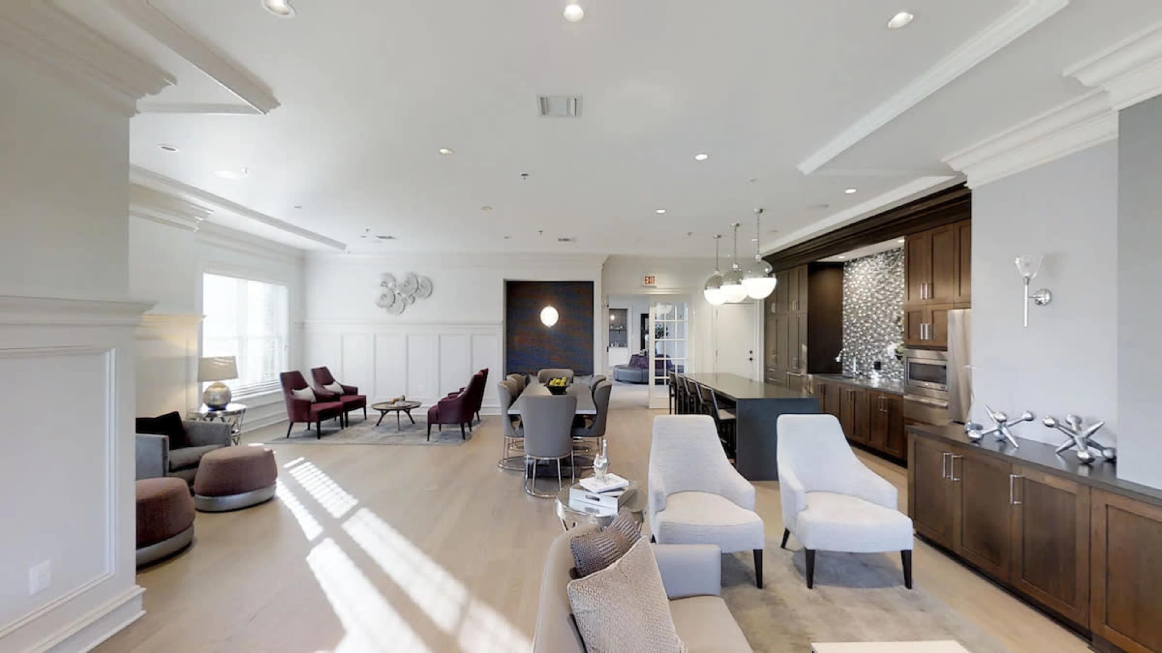 The image shows a spacious, well-lit common area furnished with a mix of chairs, tables, and decorative elements.