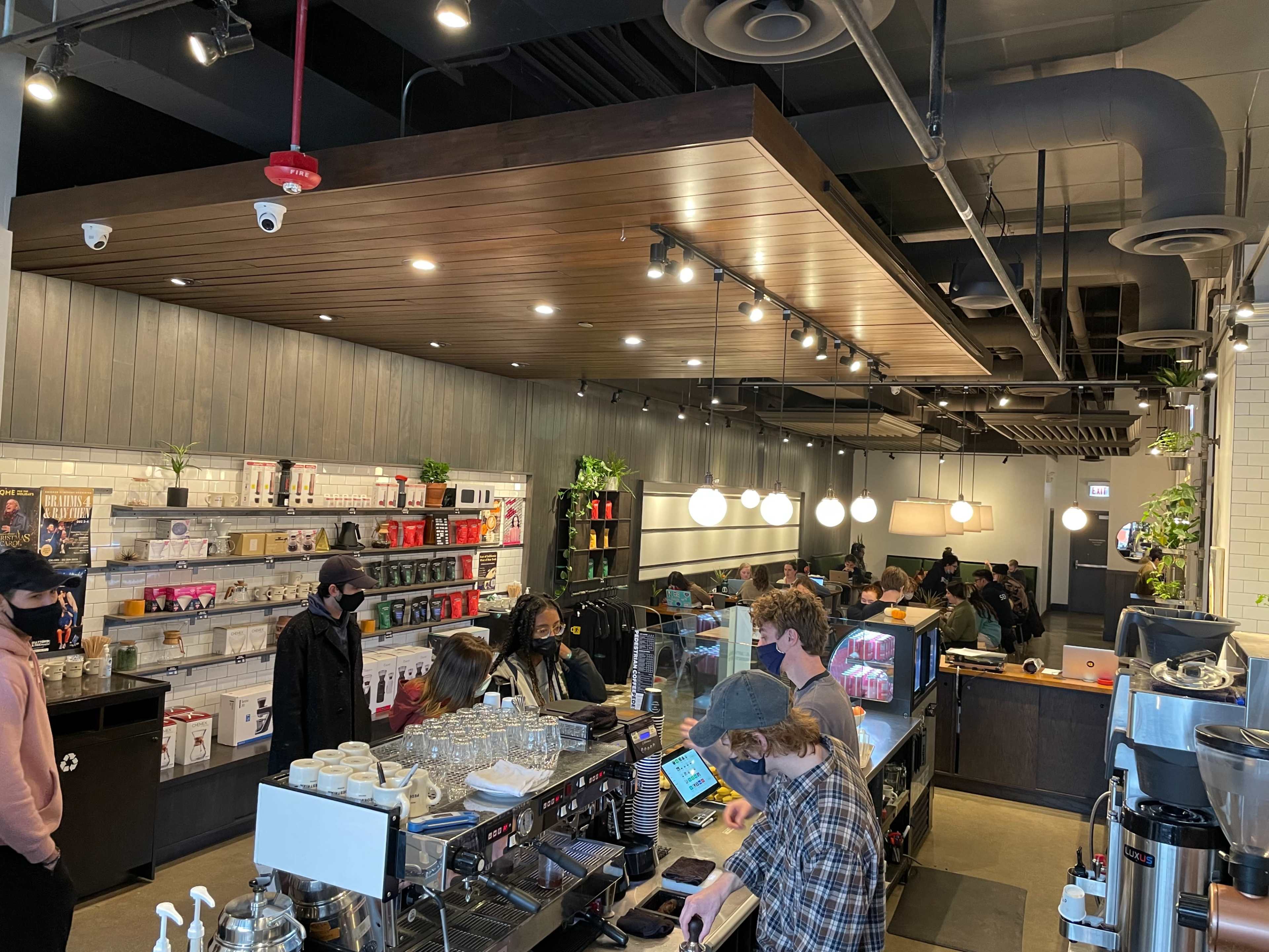 The image shows a bustling coffee shop interior with a wooden ceiling, sleek decor, and customers seated at tables while baristas serve drinks.