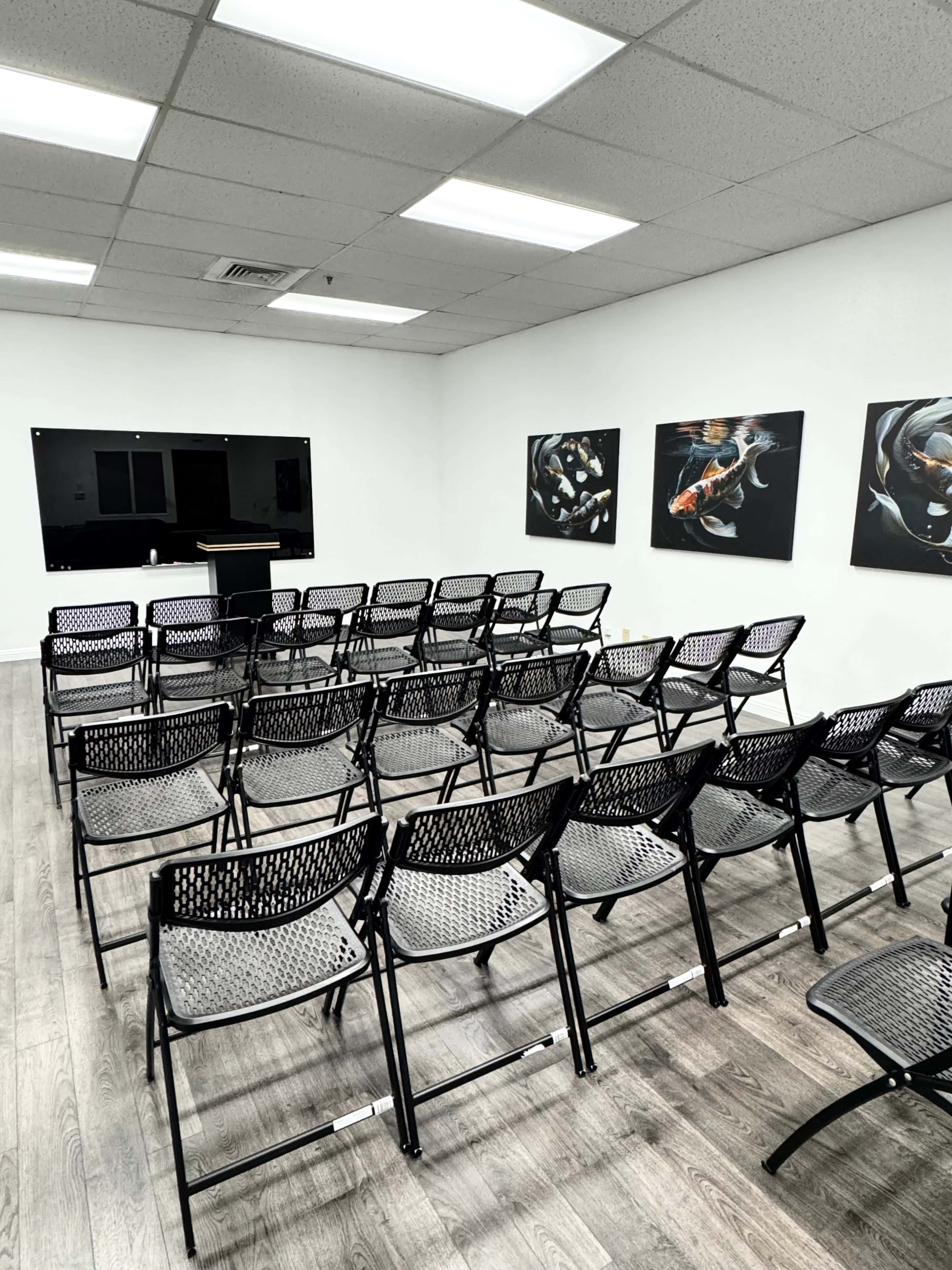 The image shows a spacious room arranged with several rows of black folding chairs facing a podium and two framed paintings on the walls.