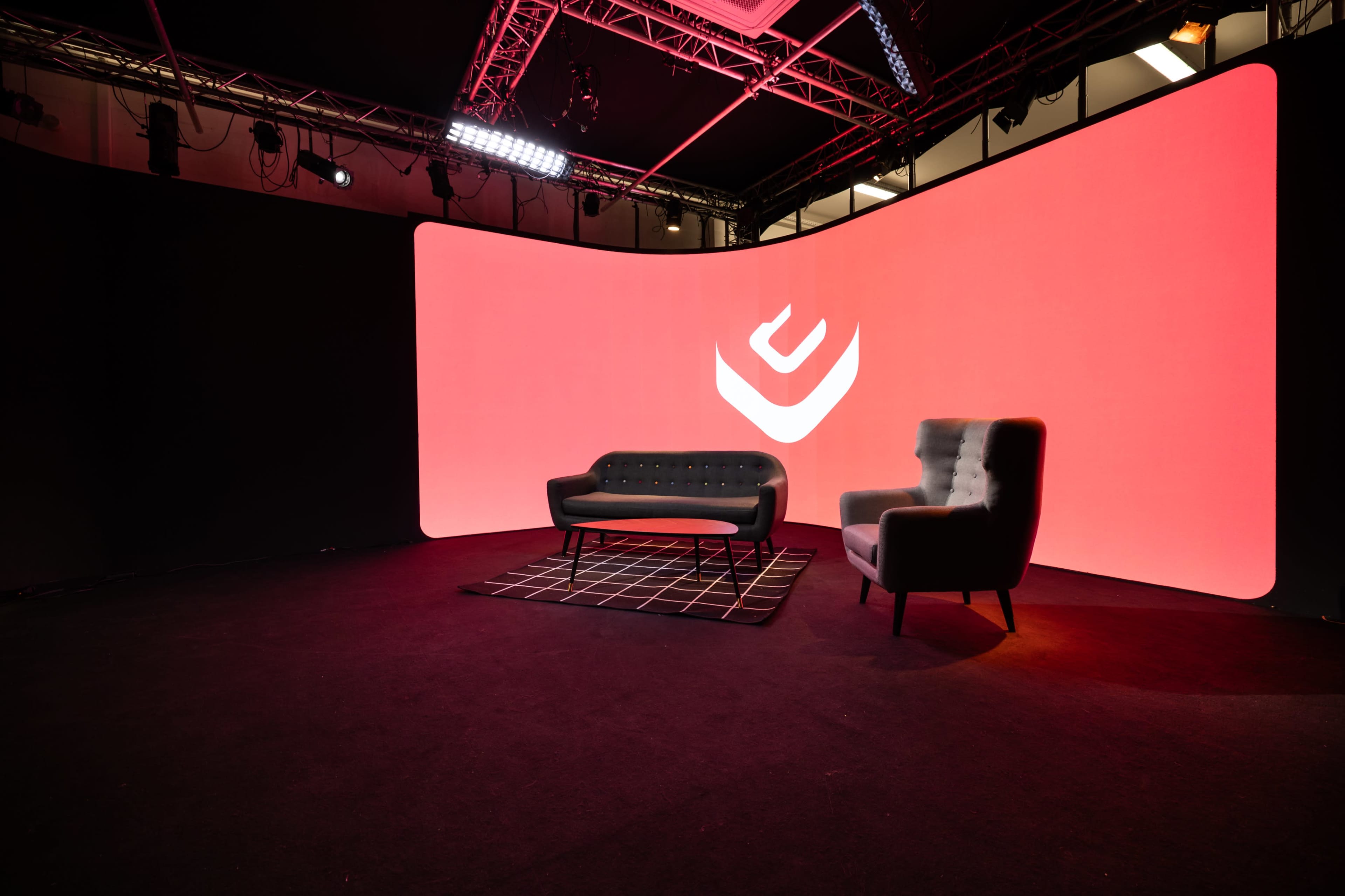 A modern stage setup featuring a black carpet, a geometric patterned rug, a black loveseat, and a gray armchair in front of a large red backdrop with a stylized logo.