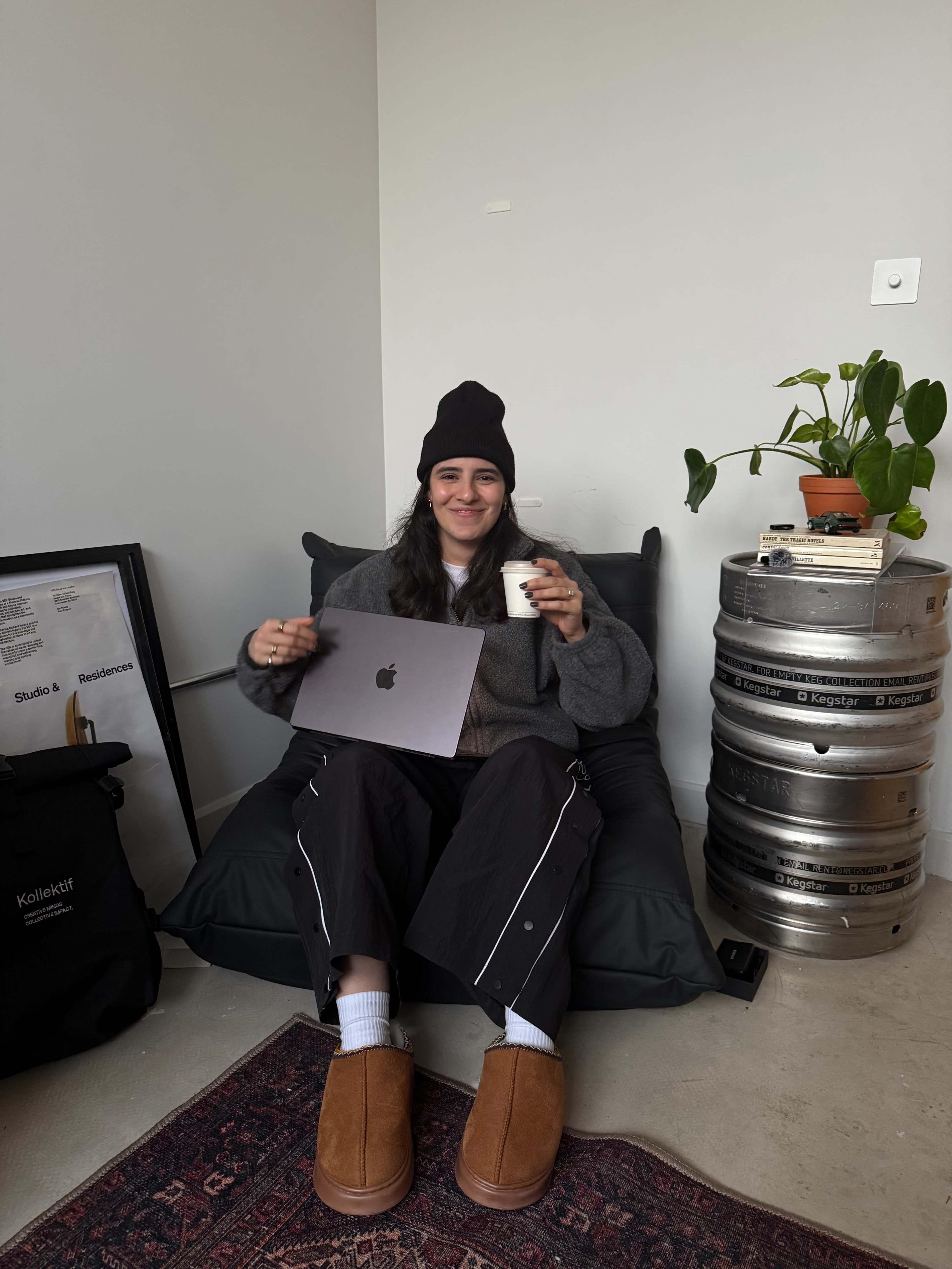 A person sitting on a beanbag chair holds a cup and a laptop, surrounded by a plant and metallic kegs.