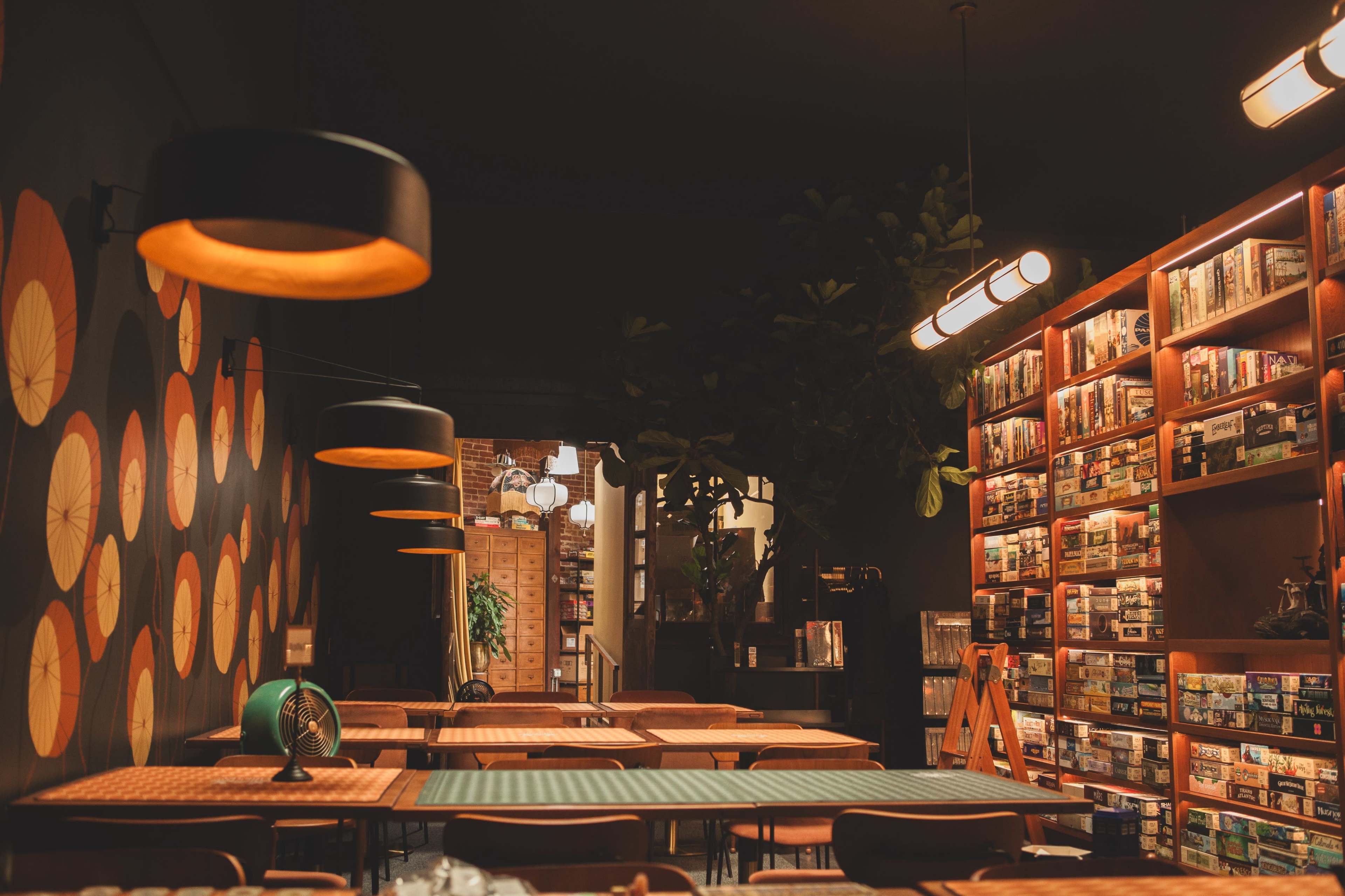 The image shows a cozy board game café with a wall decorated in vibrant circles, shelves filled with games, and wooden tables under soft lighting.