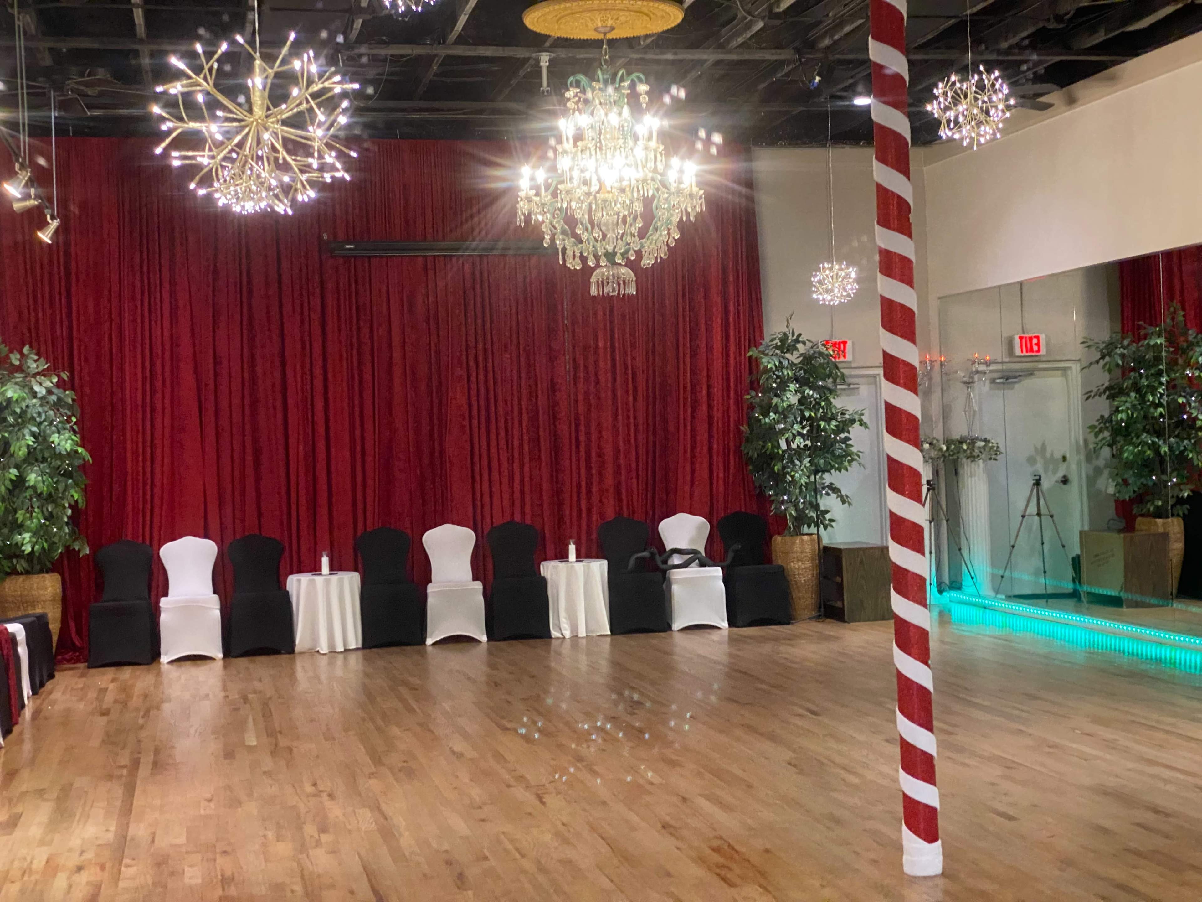 A spacious dance studio with a wooden floor, red curtains, decorative chandeliers, and seating along the sides.