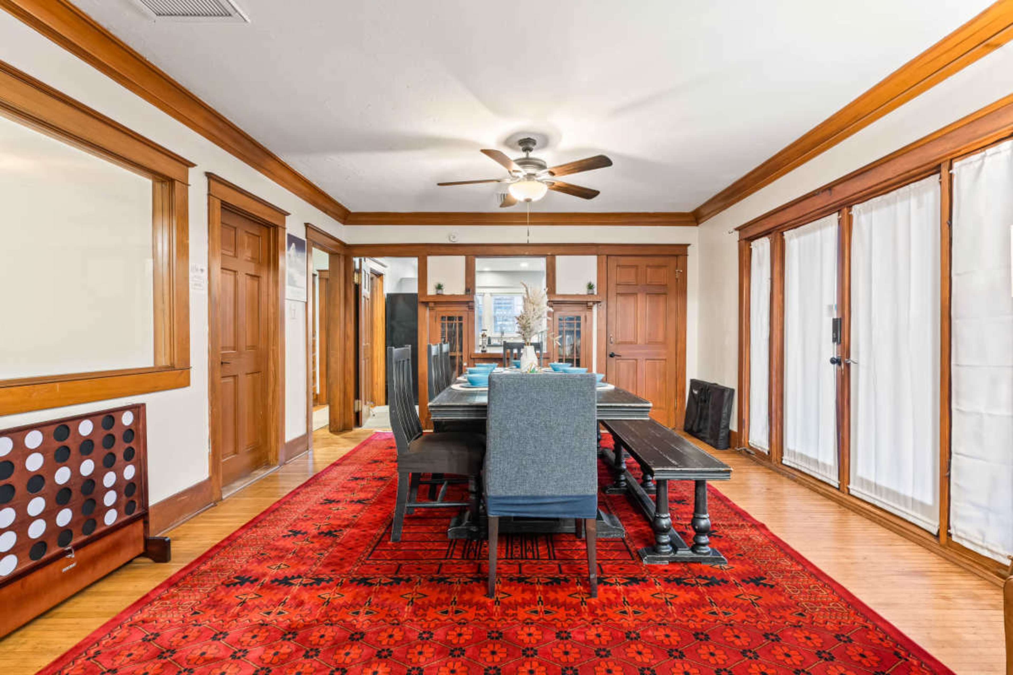 The image shows a dining room with a long table set for a meal, surrounded by chairs, hardwood floors, and a decorative red rug.