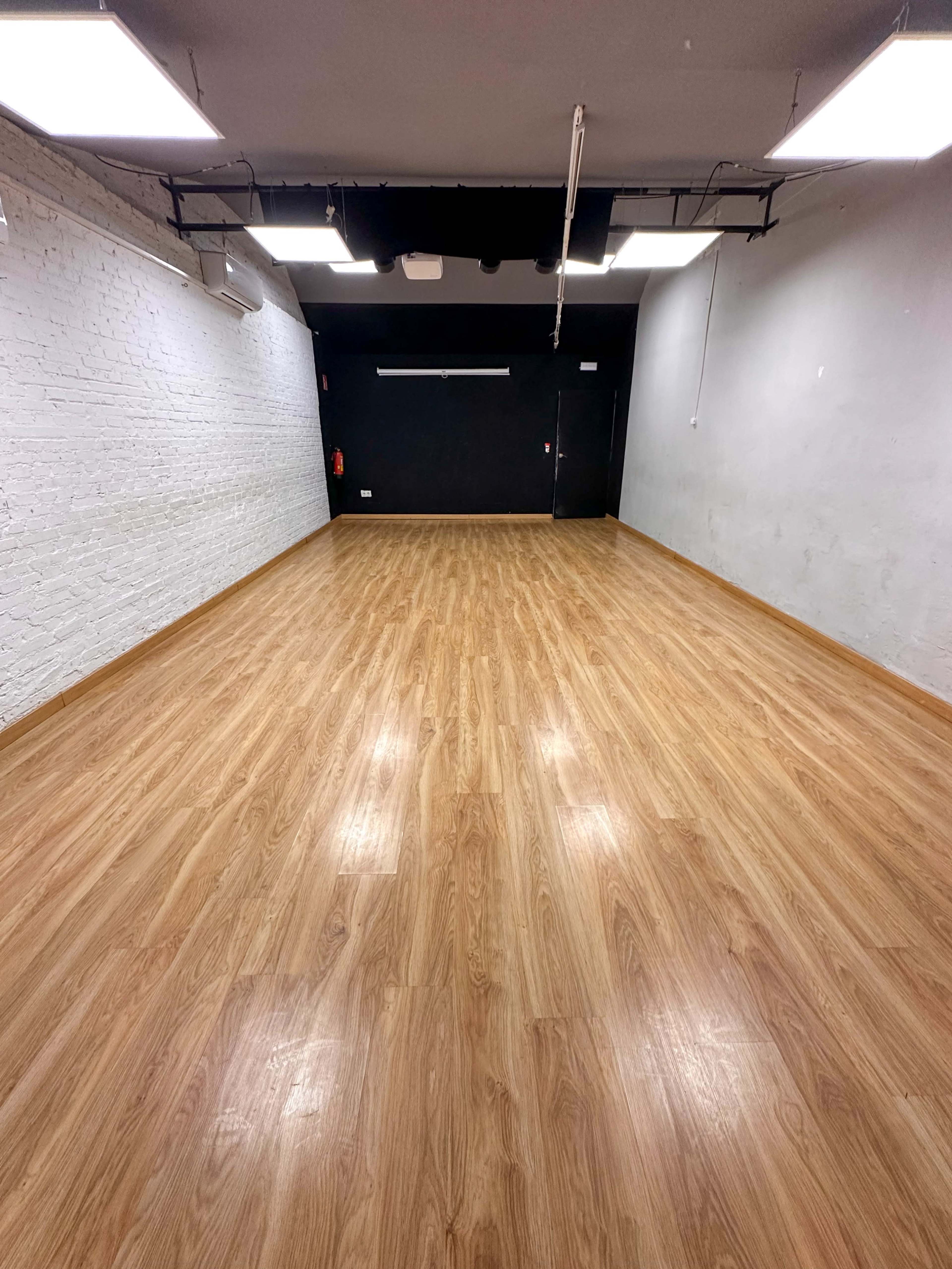 The image shows a spacious, empty room with a wooden floor, white brick walls on one side, and a dark wall at the far end.