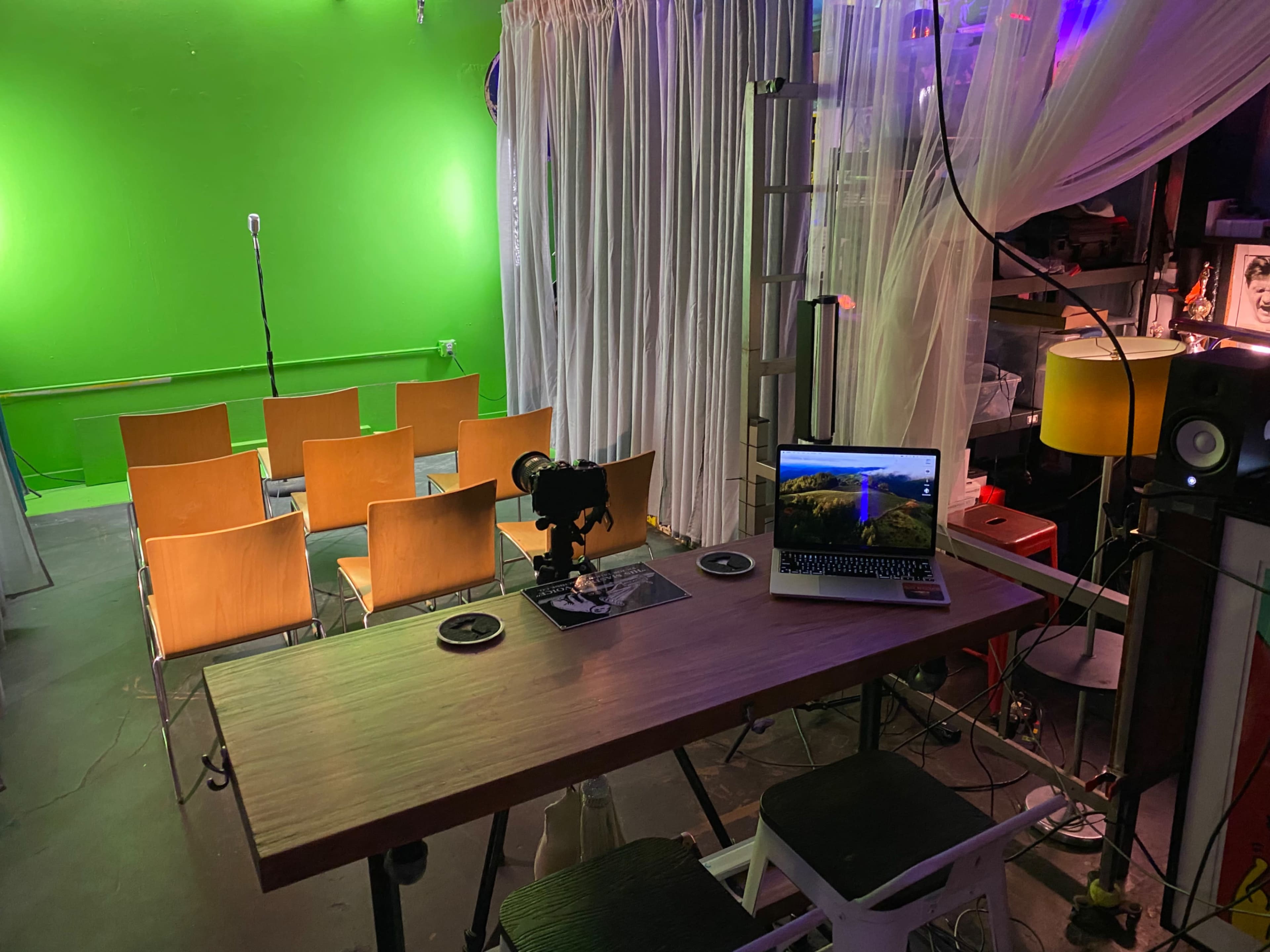 A small performance space features a green screen, rows of wooden chairs, and a table with a laptop and camera setup.