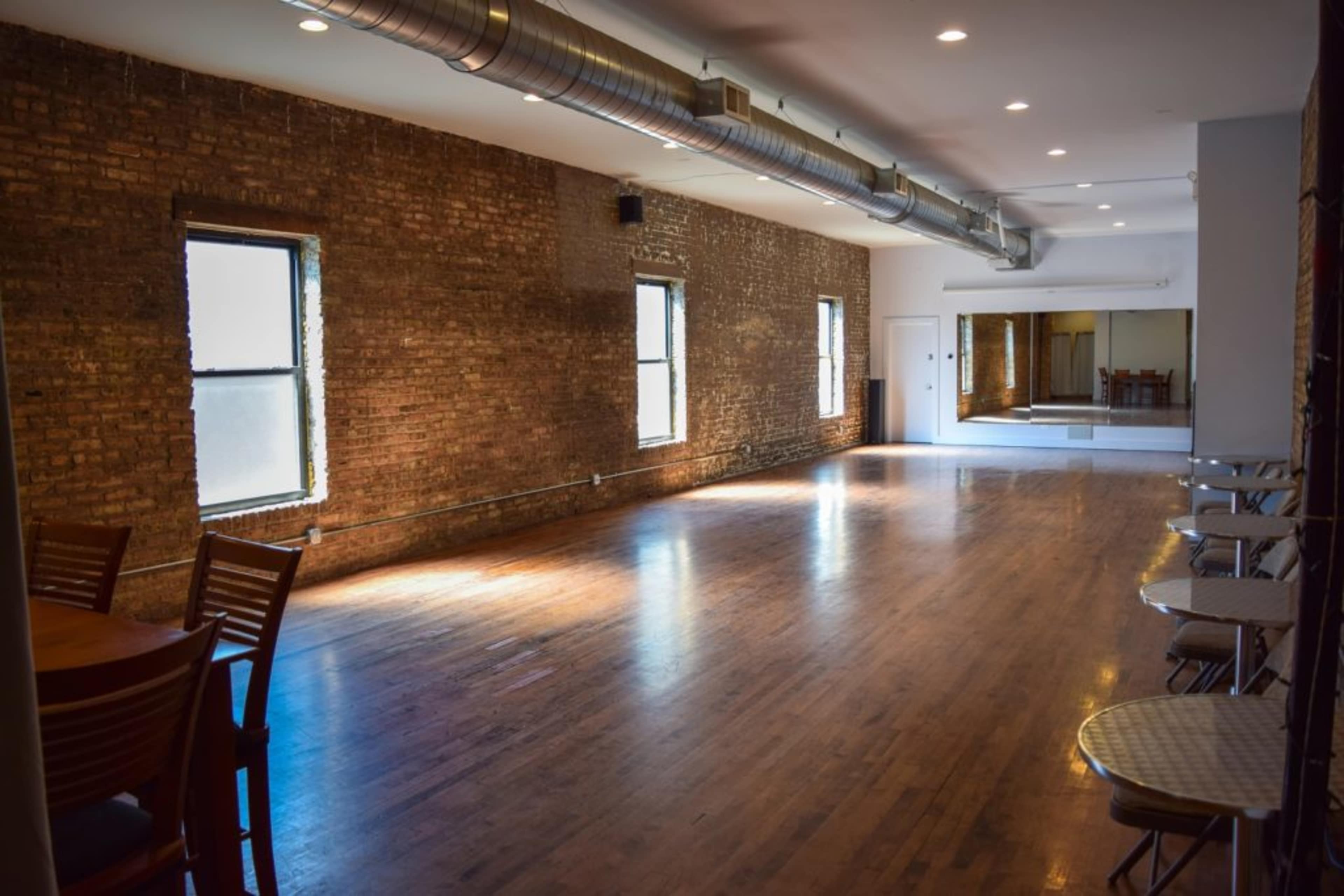 The image shows a spacious, empty room with exposed brick walls, large windows, and a wooden floor, featuring a mirrored wall at one end.