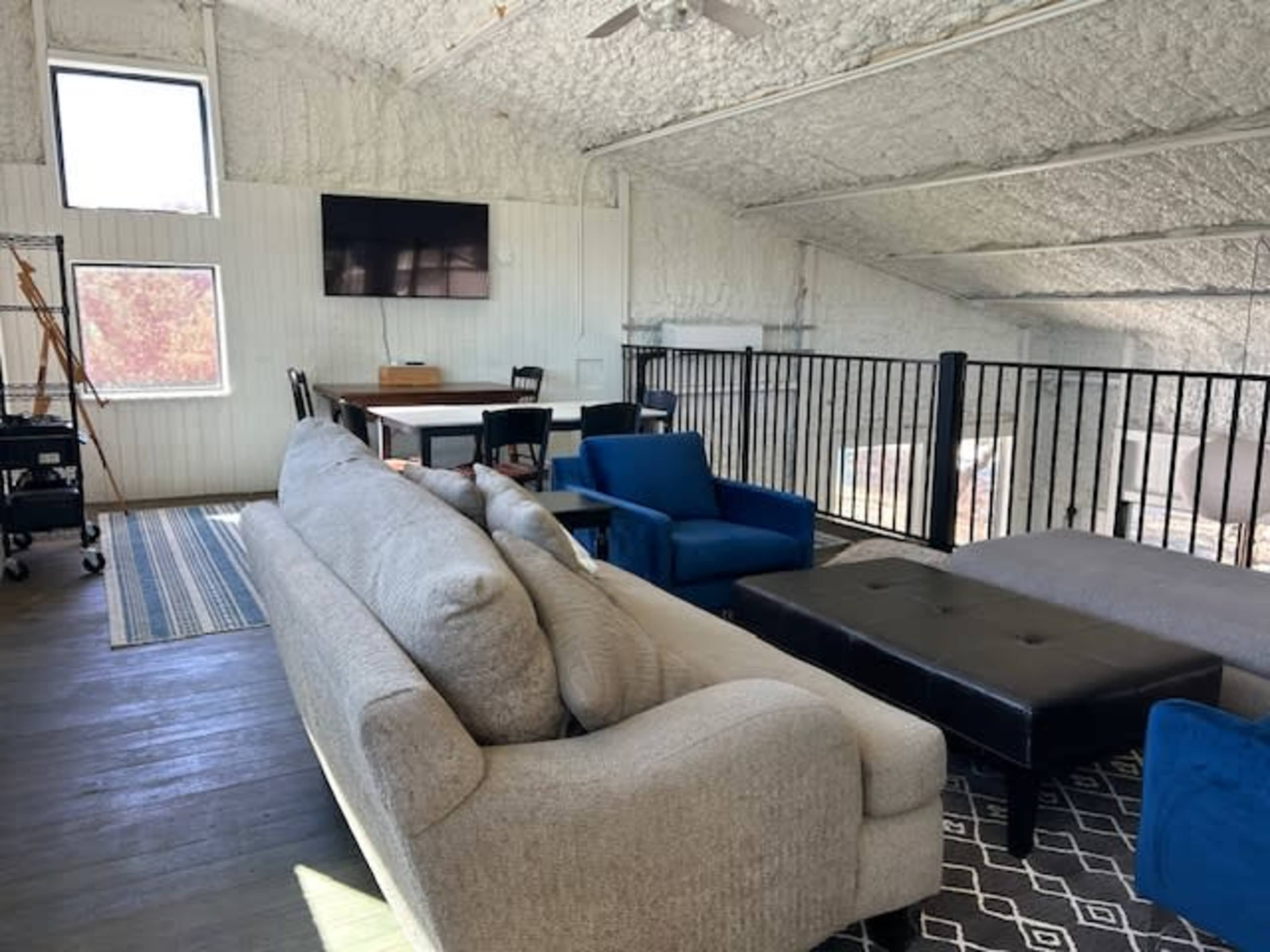 The image shows a spacious, modern room with a high ceiling, equipped with a sofa, chairs, a coffee table, and a wall-mounted television, along with large windows allowing natural light.