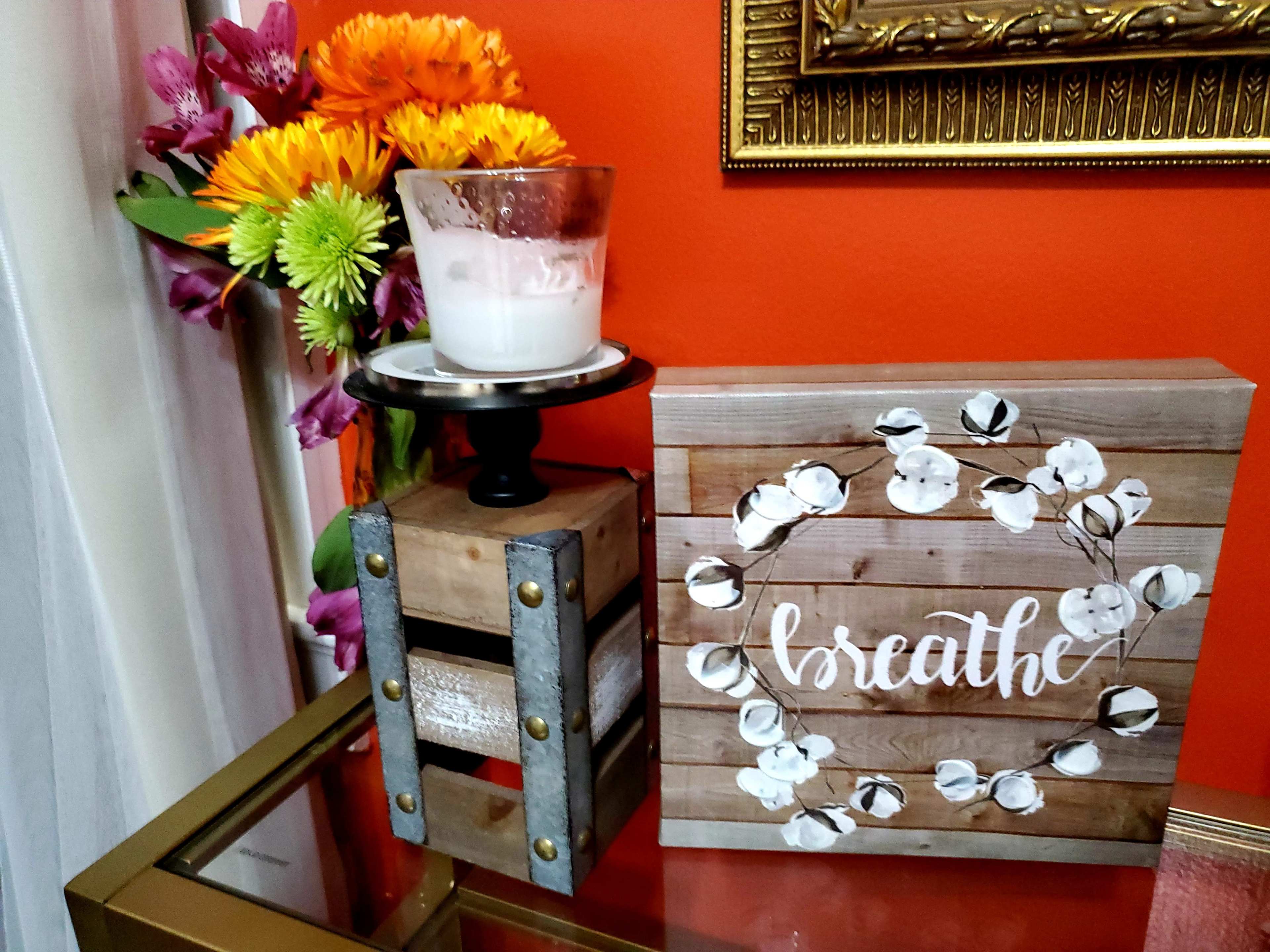 A wooden box with the word "breathe" written on it, a candle in a glass holder, and a bouquet of colorful flowers arranged nearby, all set against an orange wall.