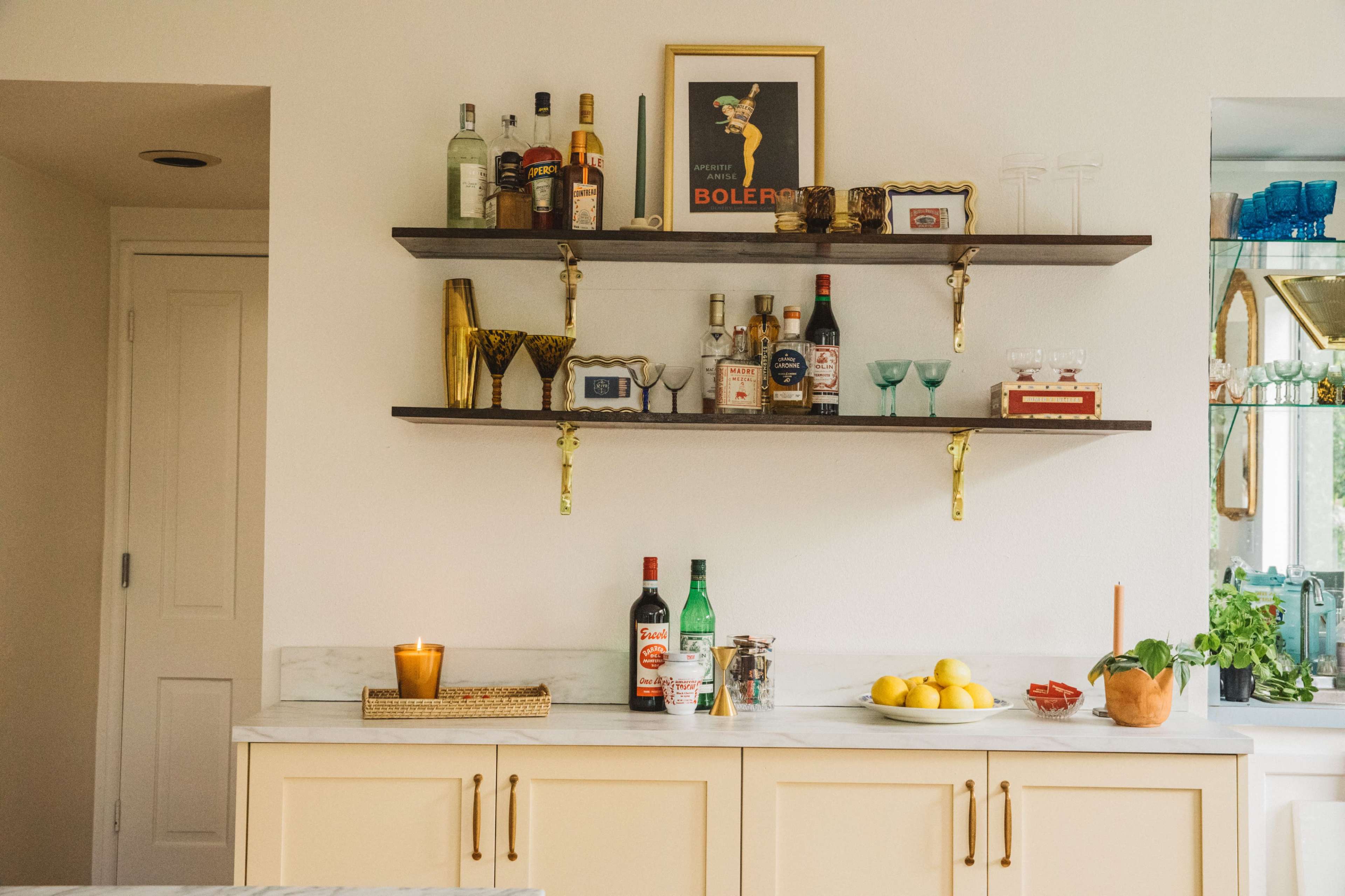 The image shows a kitchen countertop with a shelf displaying various bottles of liquor, glassware, and a candle, along with a bowl of lemons and a potted plant.