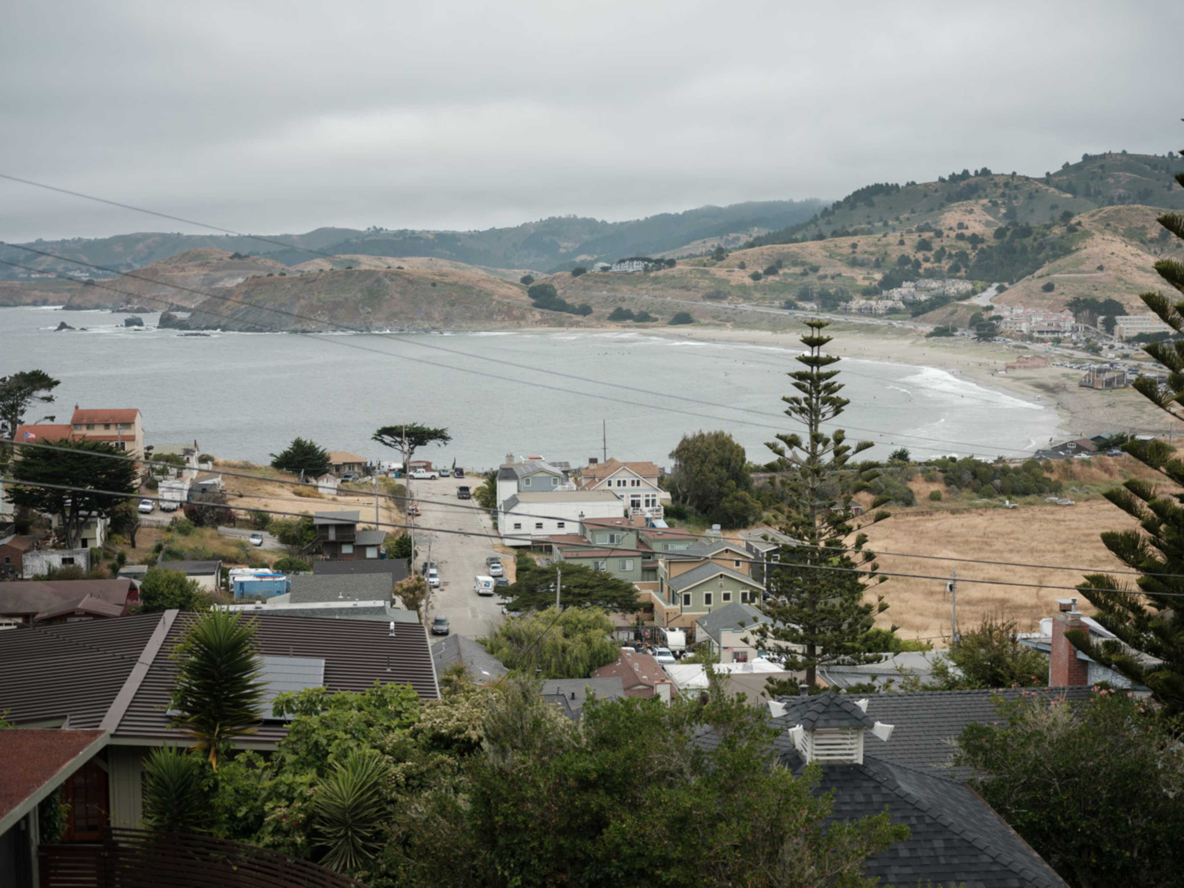A coastal landscape with a view of a beach, nearby houses, and rolling hills under a cloudy sky.