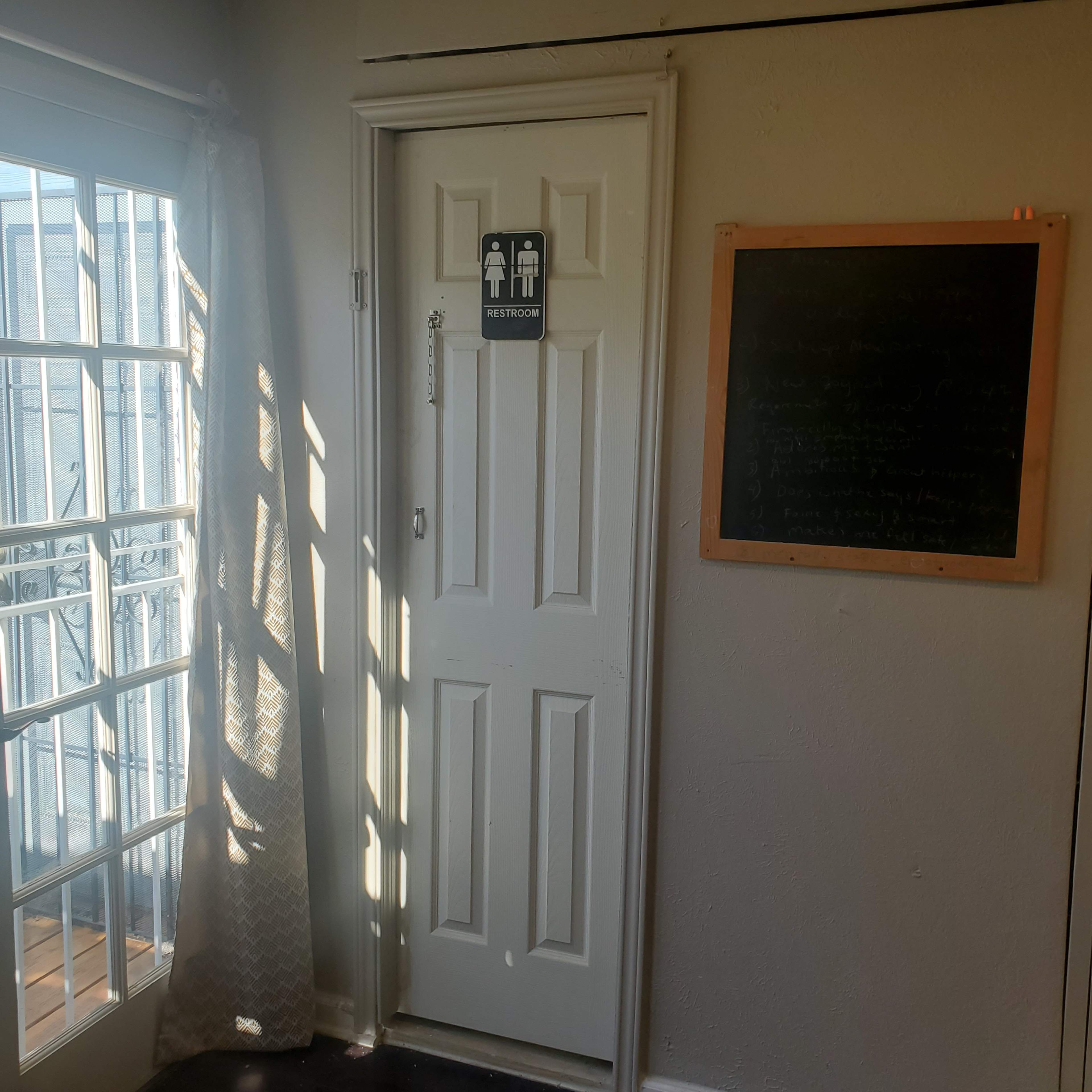 The image shows a restroom door with a gender-neutral sign, adjacent to a window with a sheer curtain and a chalkboard on the wall.
