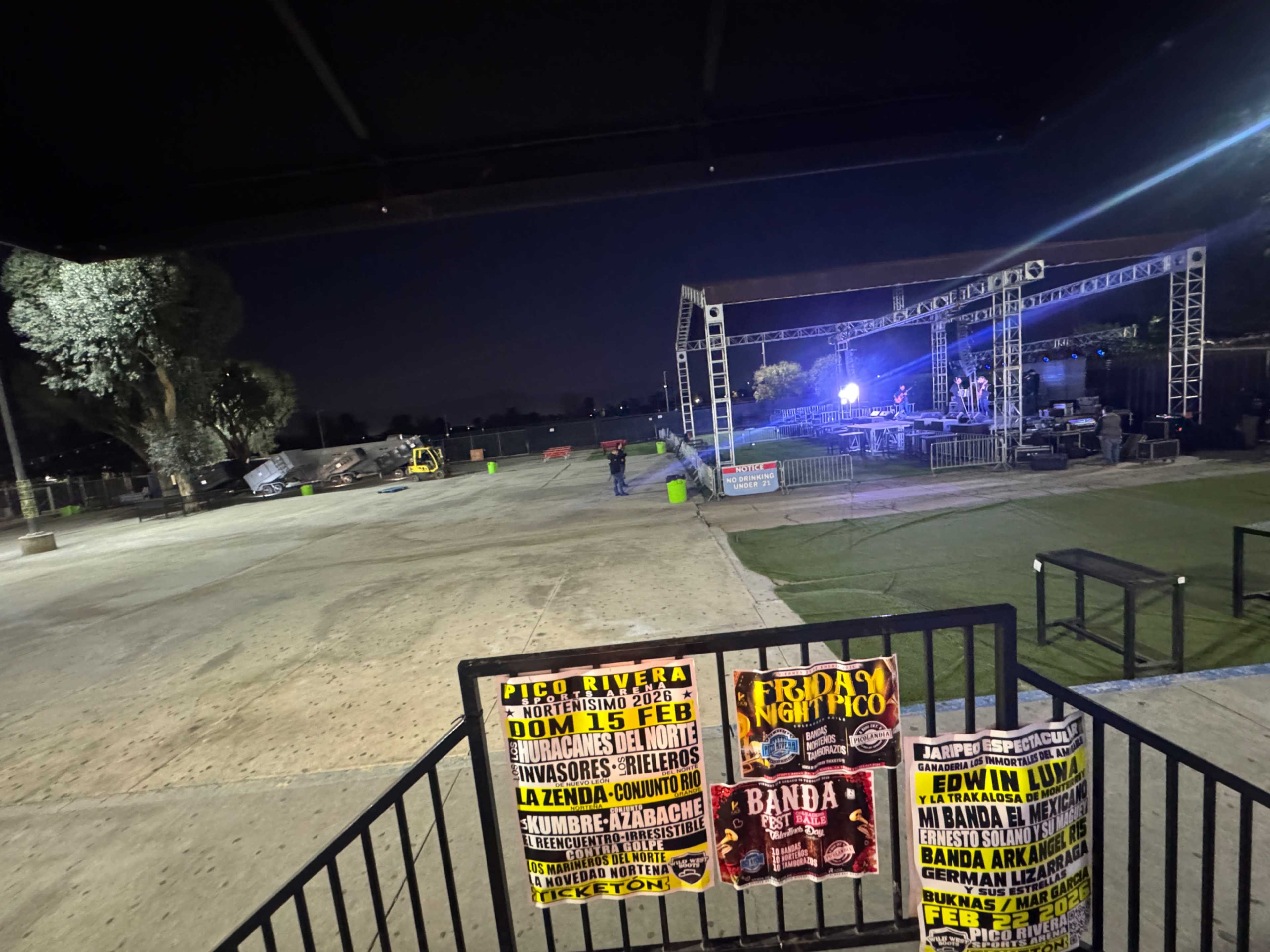 The image shows a partially set-up outdoor stage for a concert or event, with banners announcing performances and spotlights in the background.