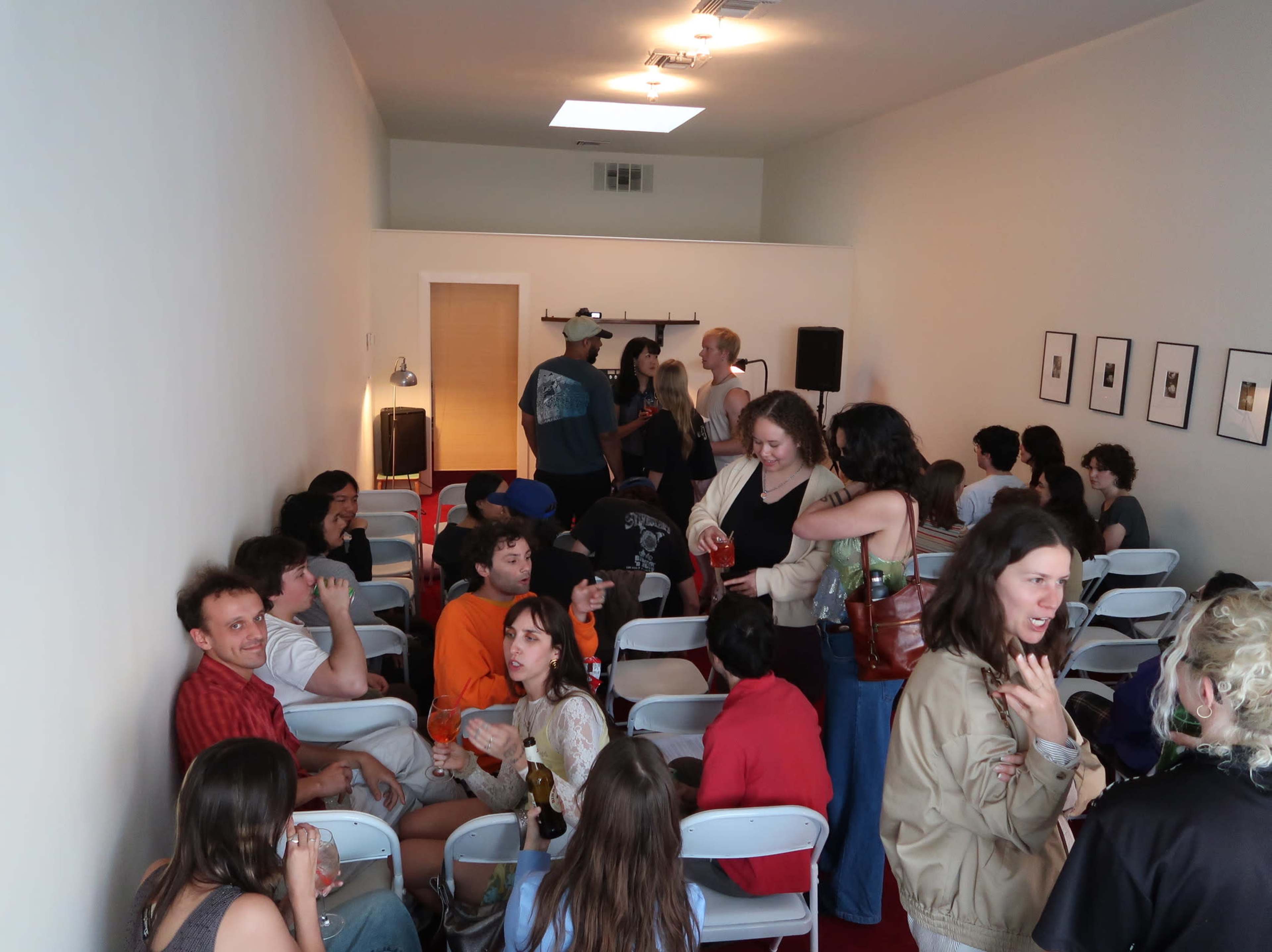 A group of people socializes in a narrow, brightly lit room, seated on white folding chairs and standing around a small stage area.