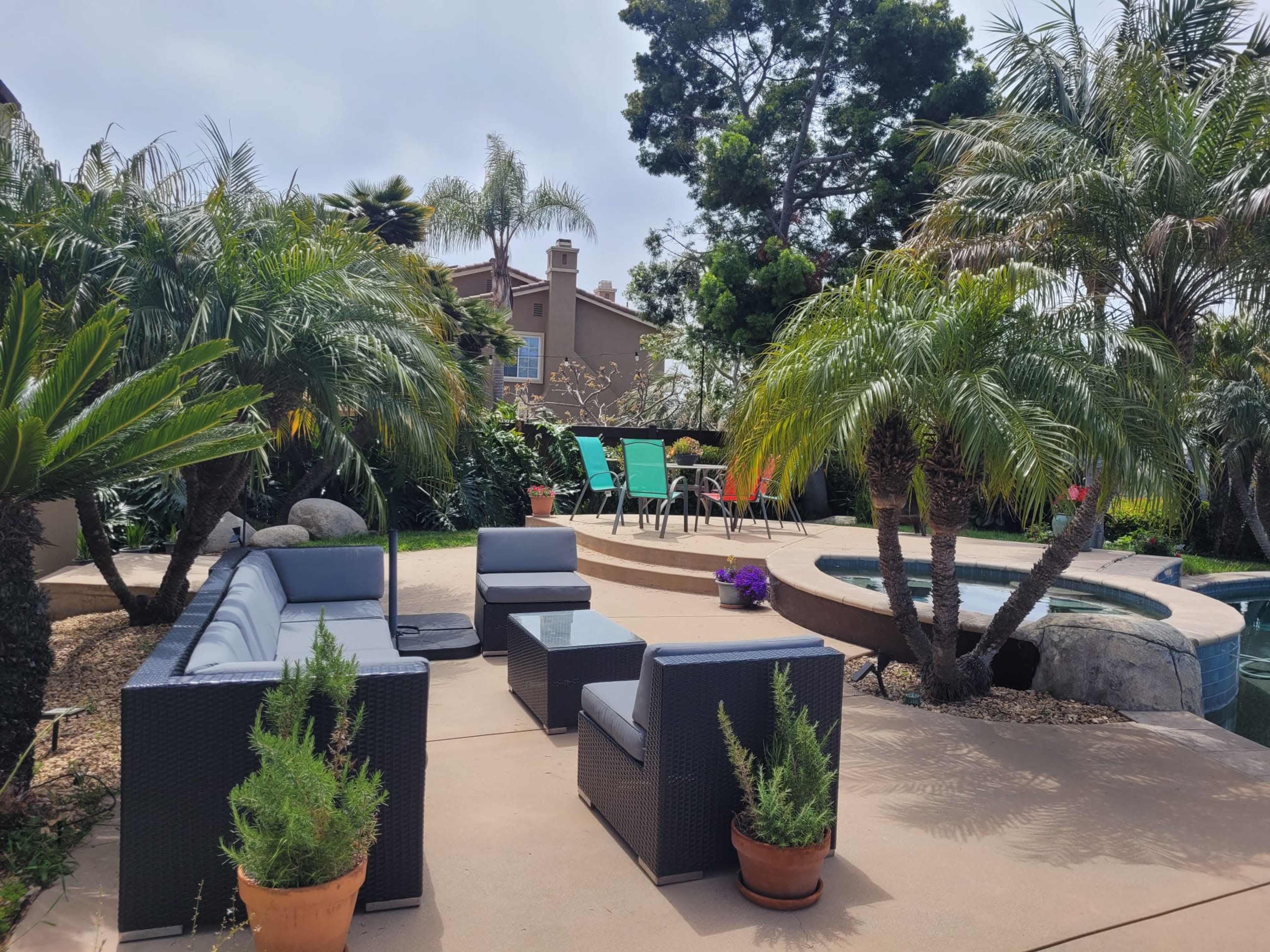 The image shows a landscaped backyard with a seating area made of dark wicker furniture, a poolside patio, and colorful chairs arranged around a table amidst palm trees.
