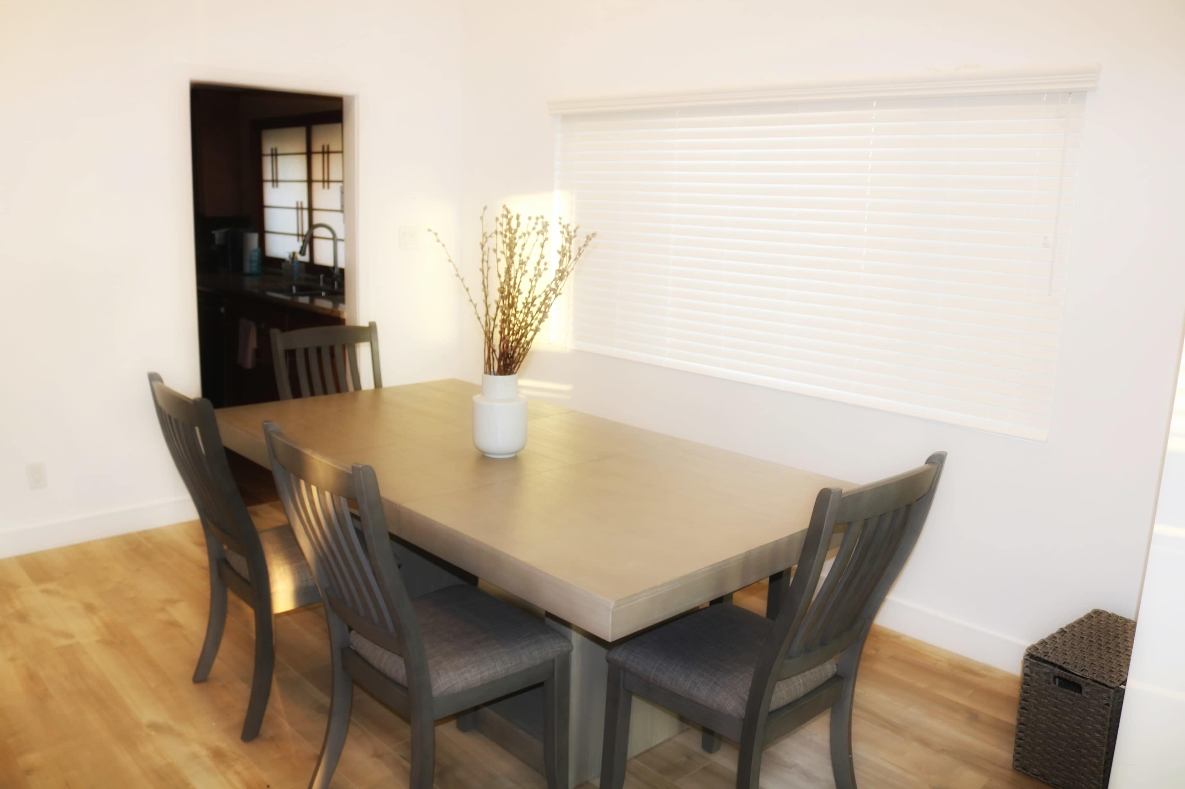 A dining table with four chairs is set in a bright room featuring a window covered with blinds and a small vase of dried flowers at the center.