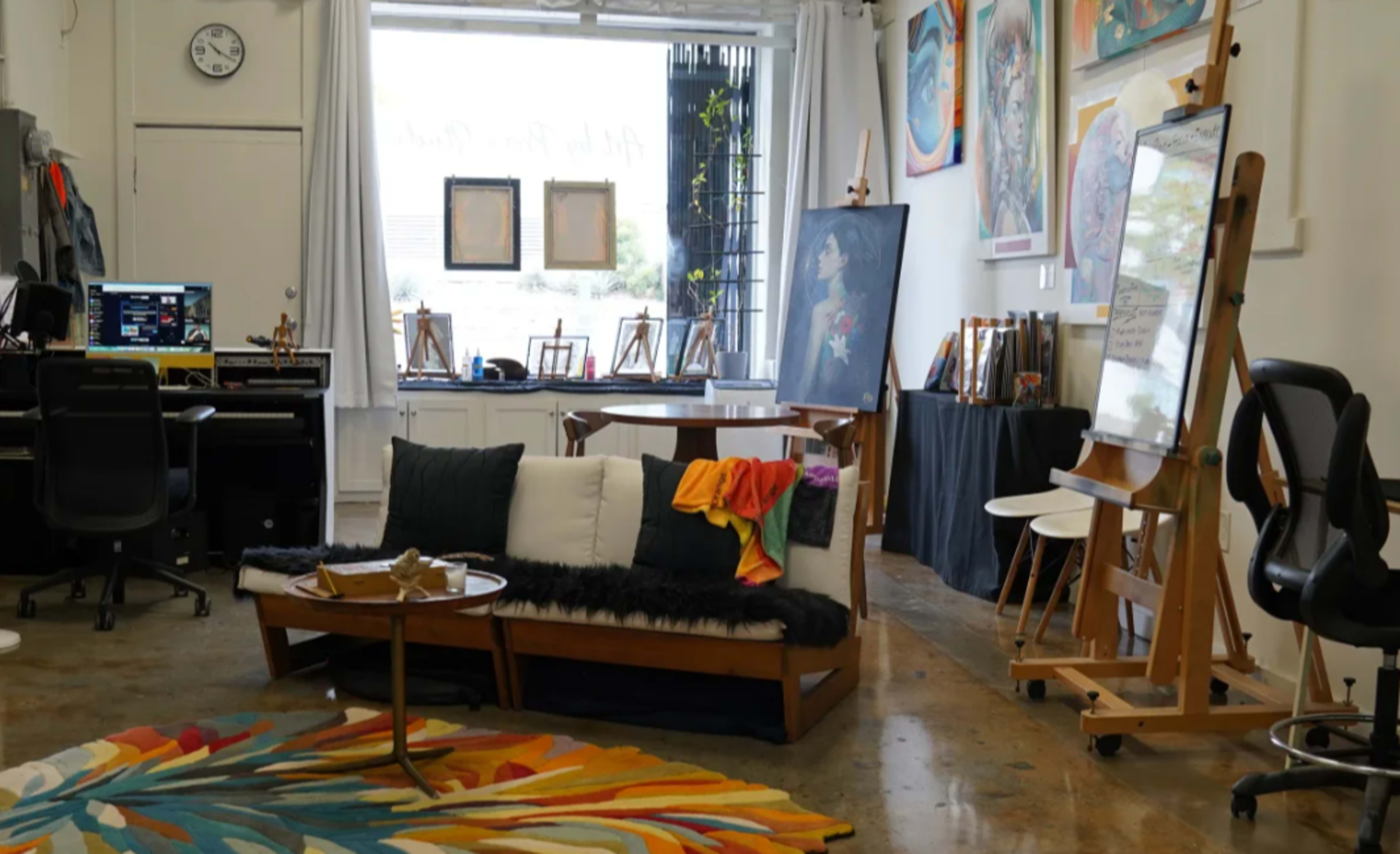 The image shows an artistic studio space with a sofa, a colorful rug, easels, and various art supplies arranged in a well-lit room.