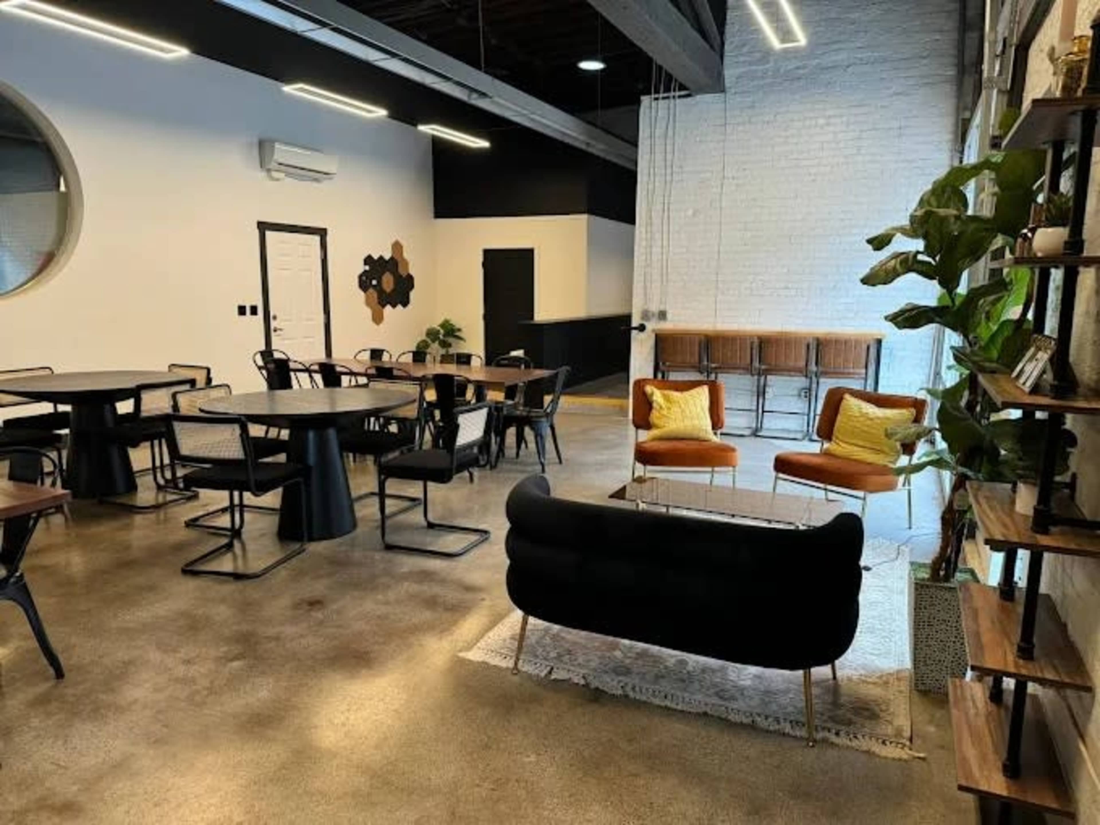 The image shows a modern, industrial-style interior with seating areas, tables, and decorative plants.