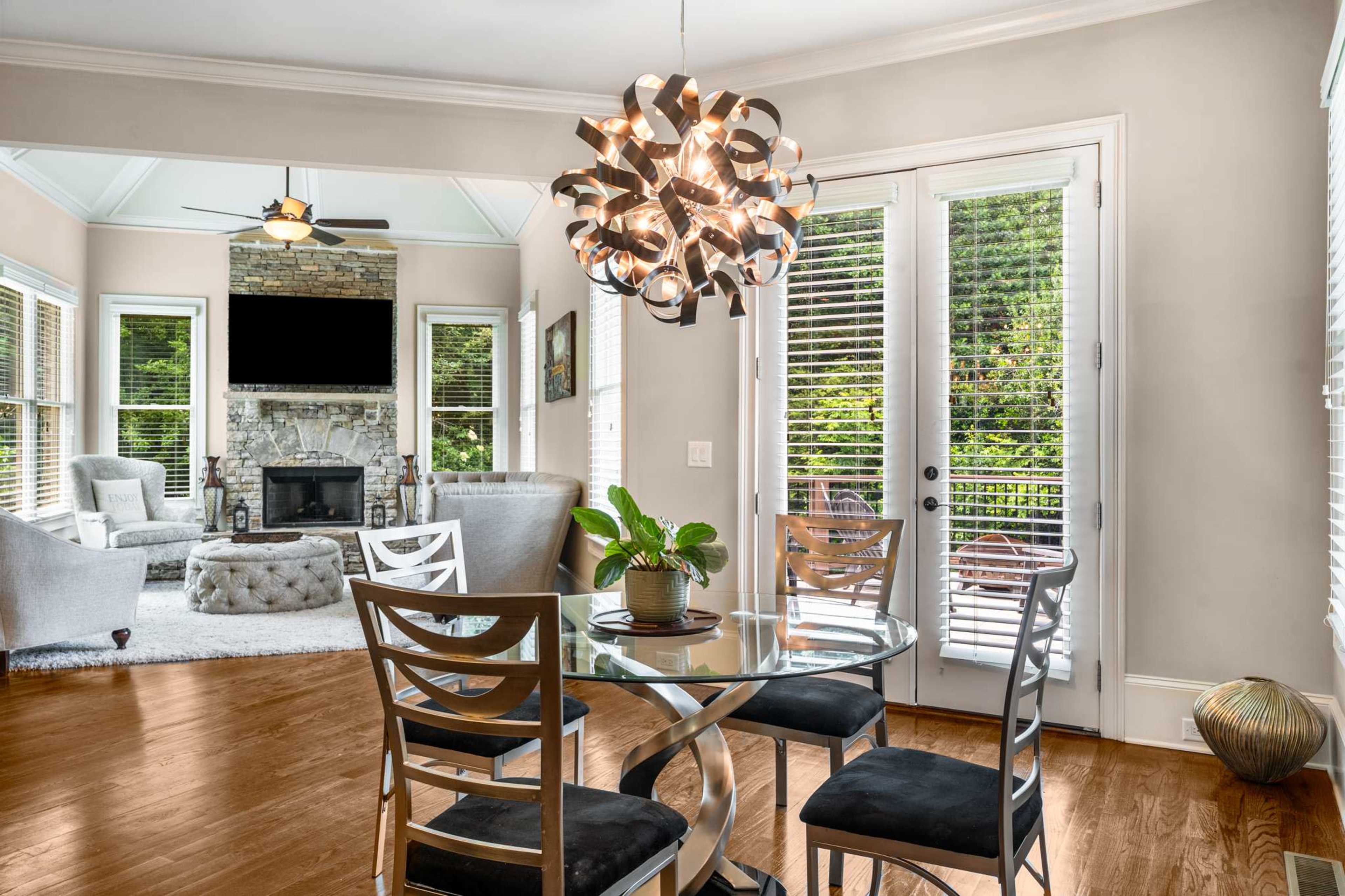 A dining area features a round glass table surrounded by four metal chairs, with a modern light fixture above, leading to a cozy living room with a stone fireplace and large windows.
