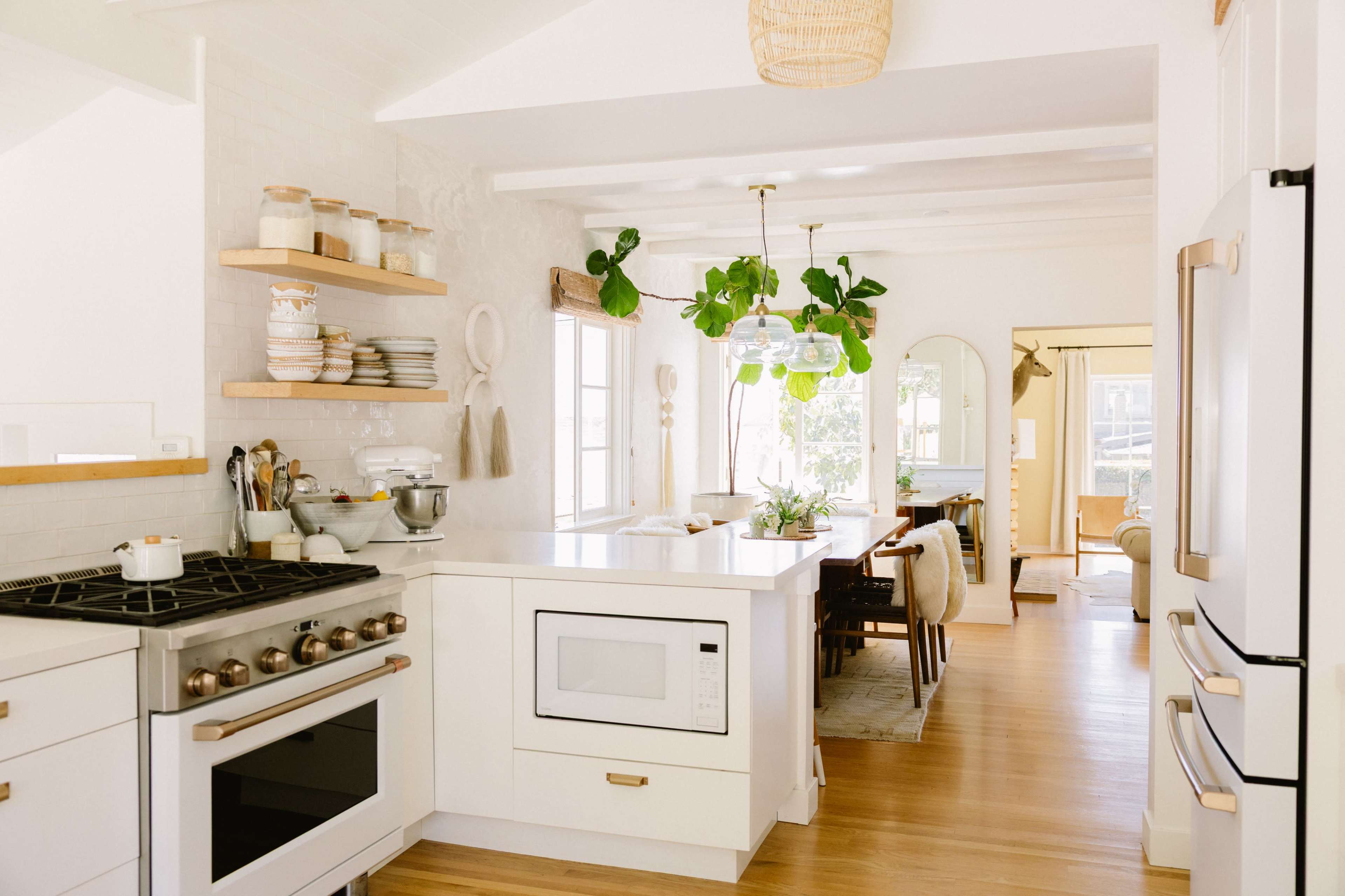 The image shows a modern kitchen with white cabinetry, a large island, and open shelving displaying kitchenware, leading into a dining area visible at the far end.
